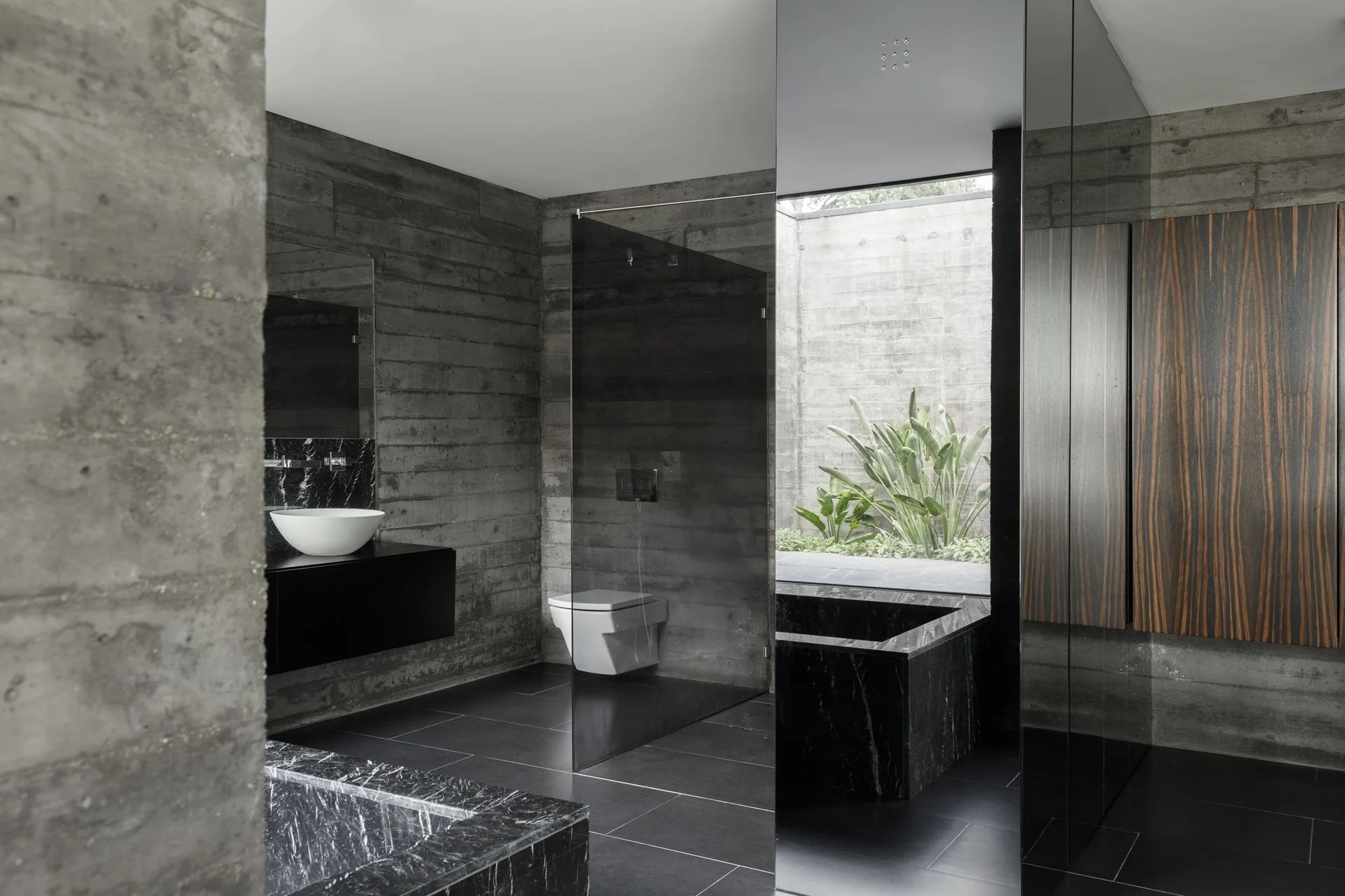 A minimalist bathroom with board-formed concrete walls, a black marble tub, a floating vanity, and a window displaying lush green plants.