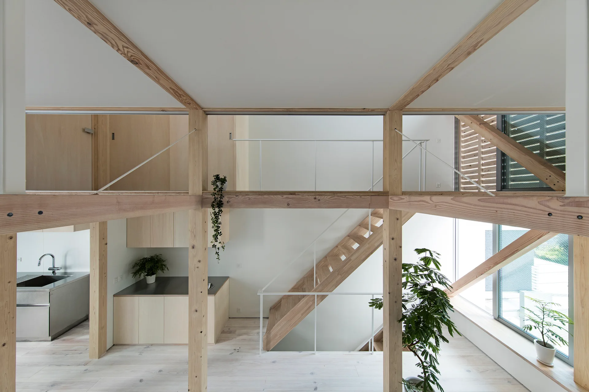 An open-plan minimalist kitchen and wooden staircase framed by exposed light wood timber posts and clean white walls.