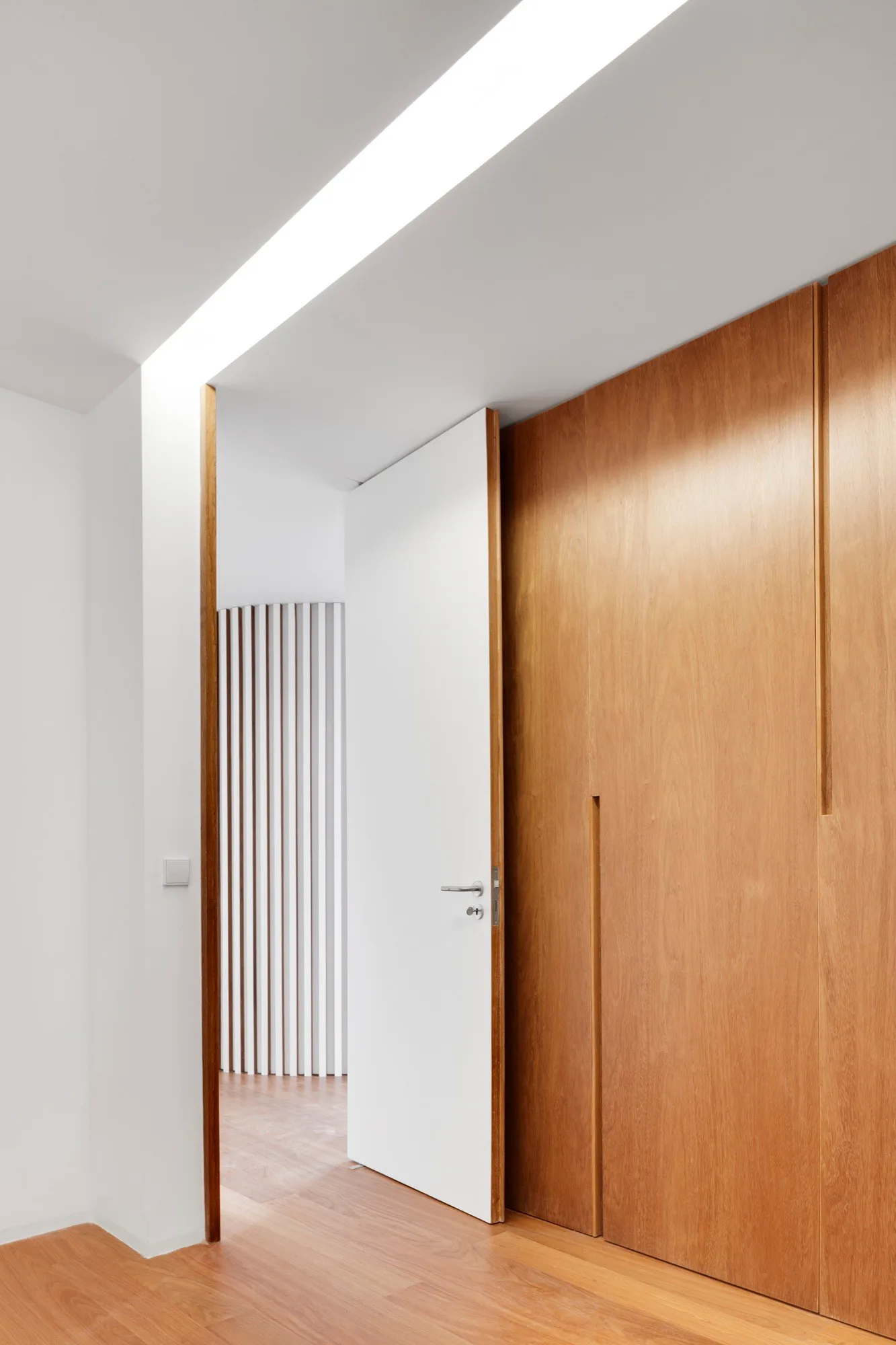 A minimalist hallway featuring a white door open next to wood-paneled walls with integrated vertical handles, and a slatted screen in the background.