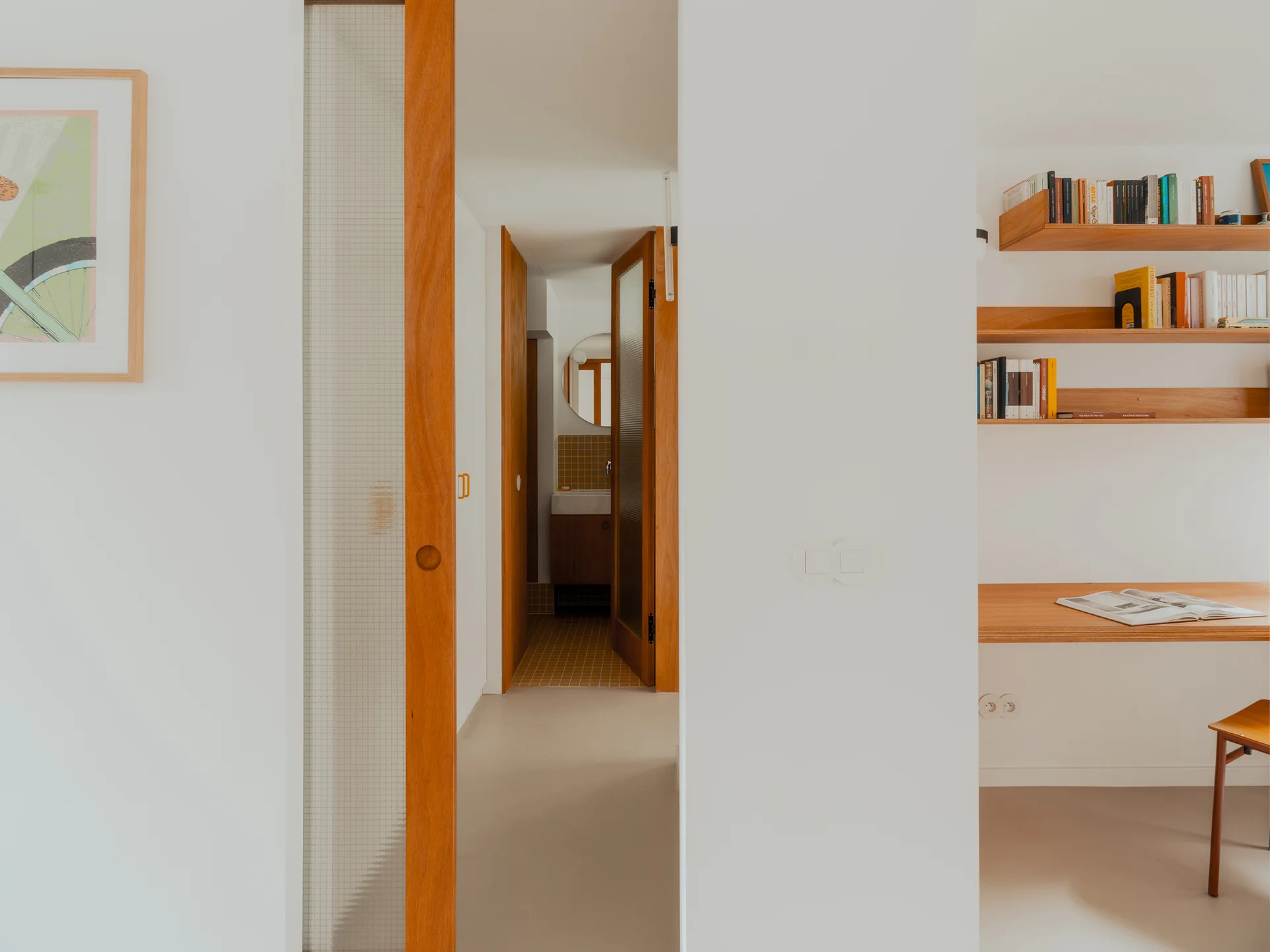 A minimalist white hallway features a built-in wooden desk with open shelves, a sliding door with translucent glass, and a view of a tiled bathroom.