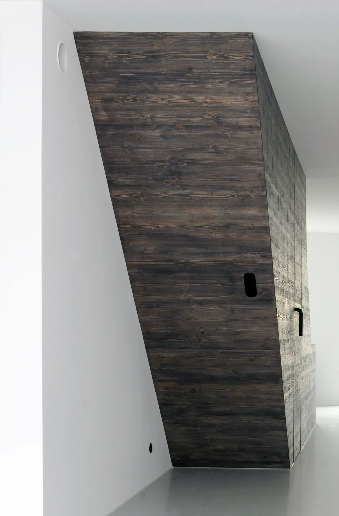A minimalist interior features a sharply slanted dark wood paneled volume next to a clean white wall and light grey floor.