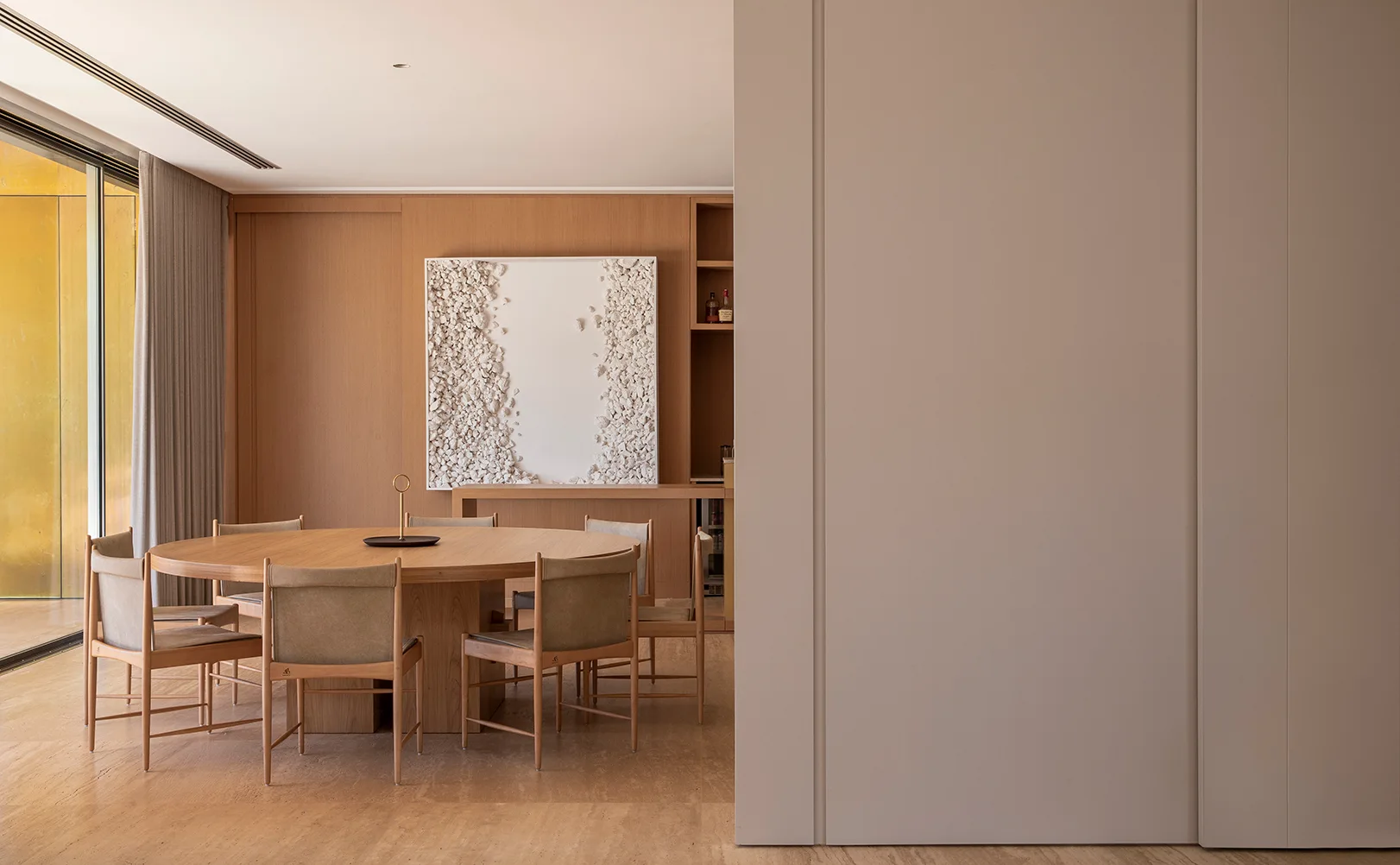 A minimalist dining room with a round wooden table, matching chairs, and textured white wall art mounted on a wood-paneled wall.