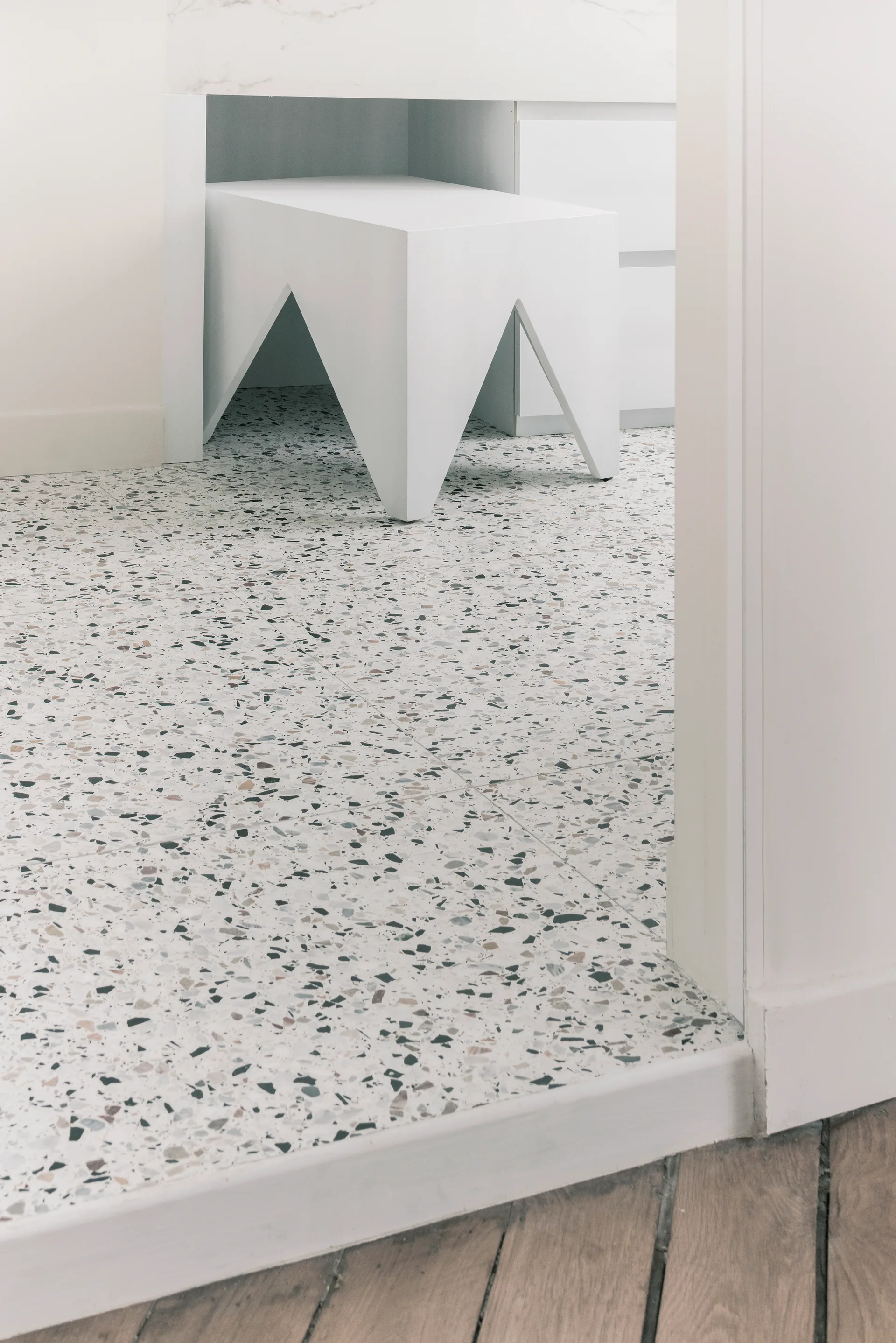 A transition over a white wood threshold from wood flooring to multi-colored flecked white terrazzo with a modern geometric stool.