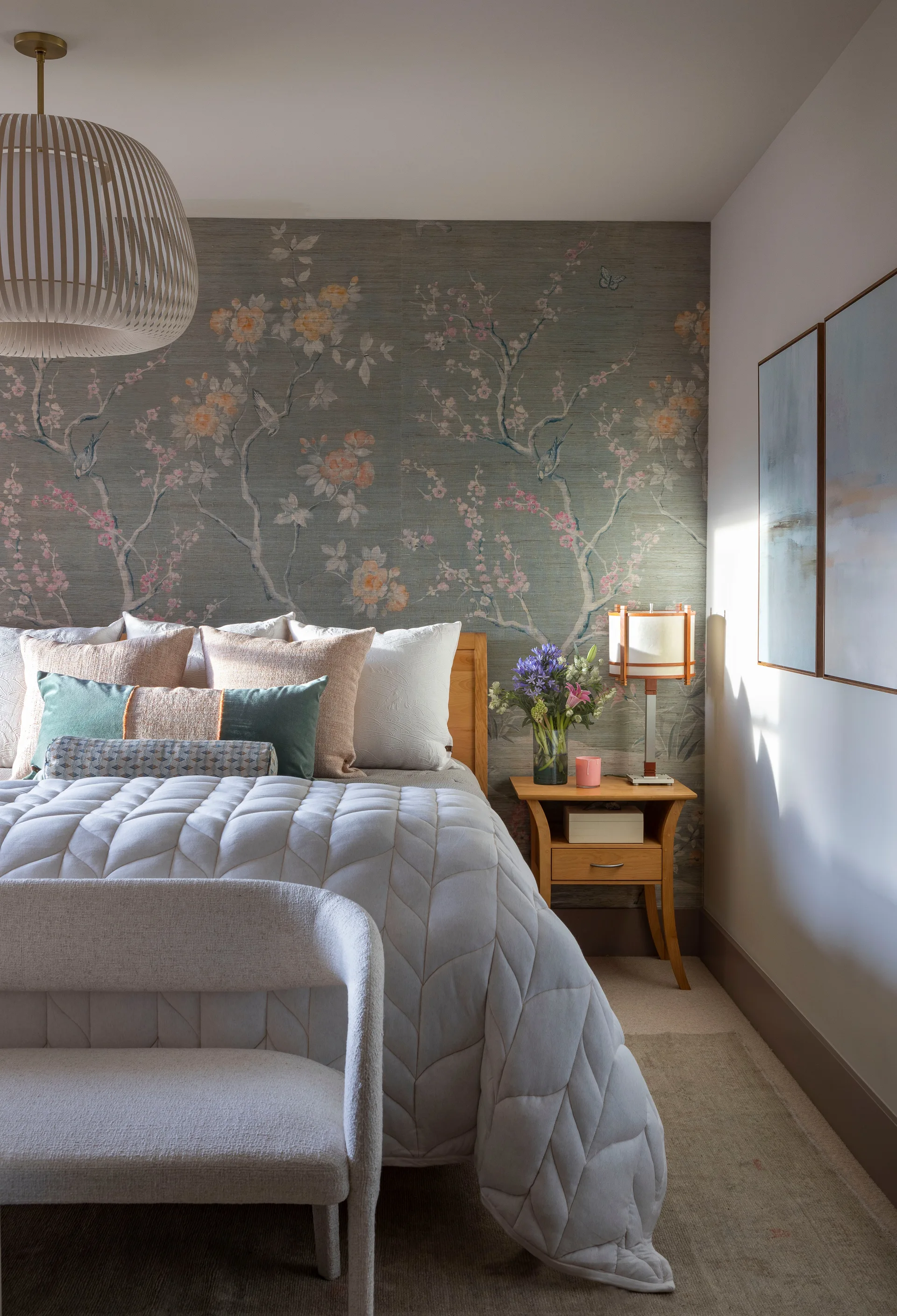 A bedroom featuring a bed with layered pillows and a quilted coverlet against a botanical wallpaper accent wall next to a wooden nightstand.