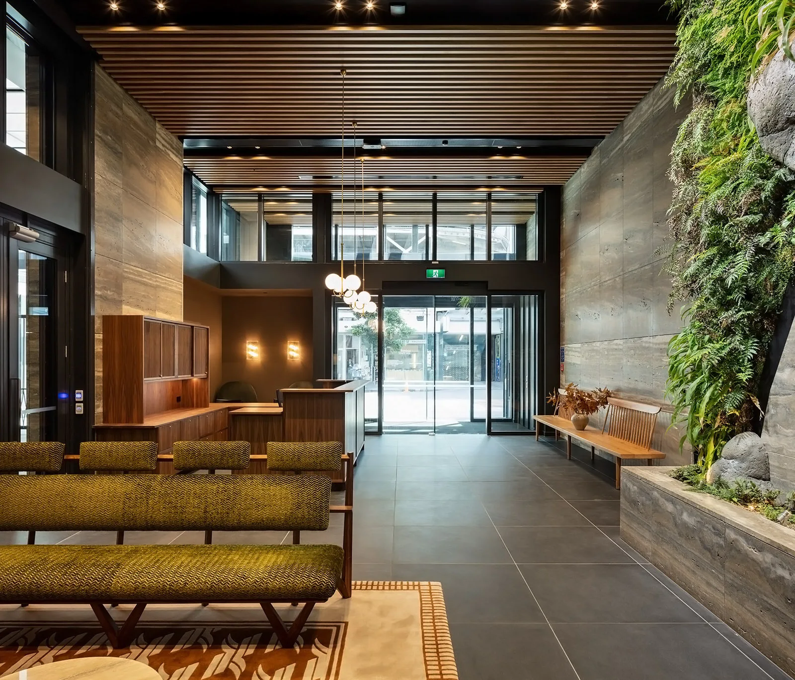 A modern lobby entryway features a slatted wood ceiling, a vertical green plant wall, and an olive green chevron patterned waiting bench.
