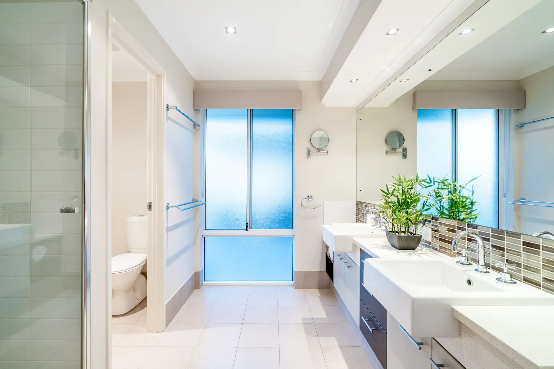 A modern bathroom showing a double sink vanity with a glass mosaic backsplash, a large wall mirror, and a frosted window.