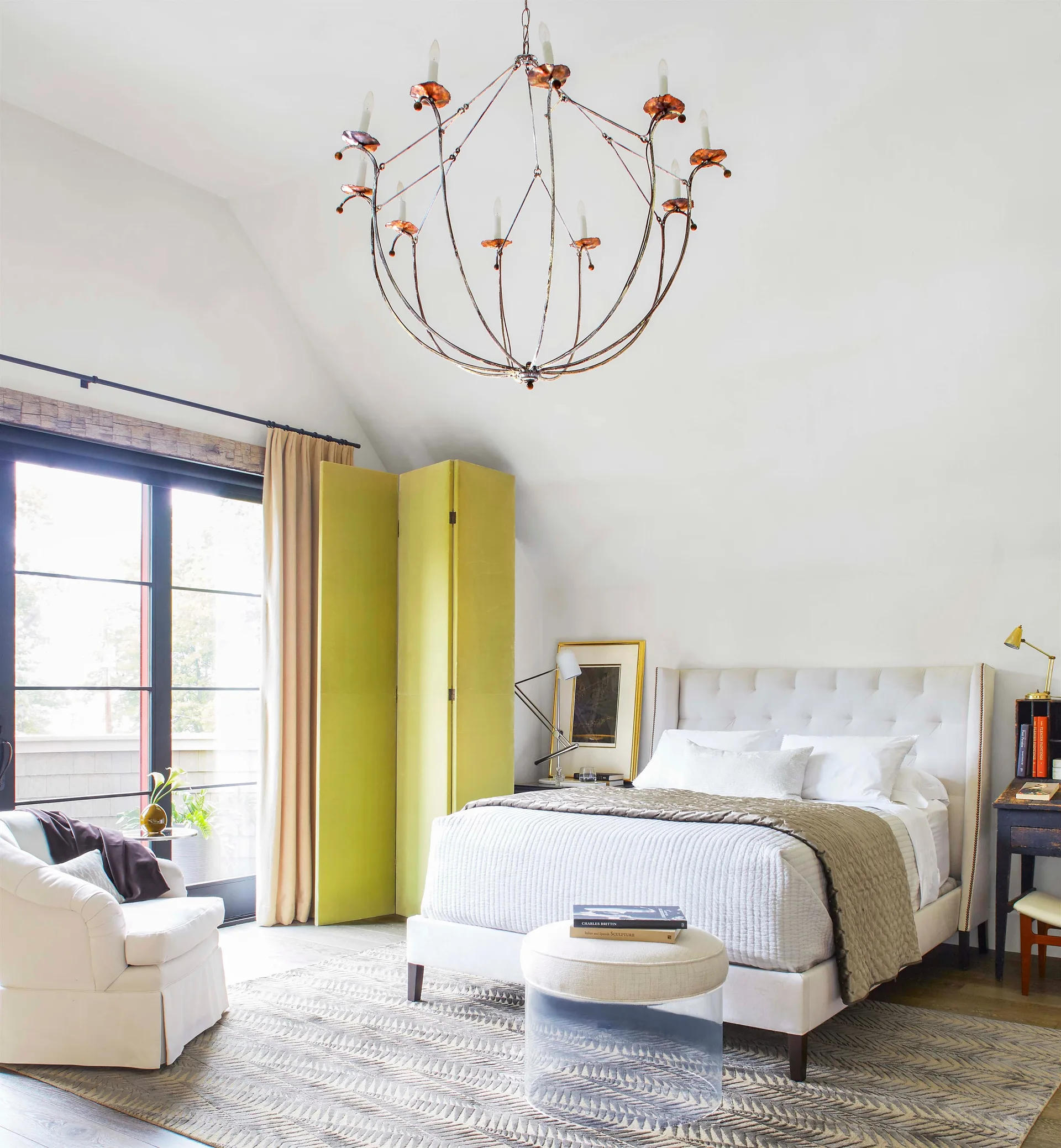 A bright bedroom with a tufted upholstered bed, a lime green folding screen, a clear acrylic ottoman, and a metal chandelier under a vaulted ceiling.