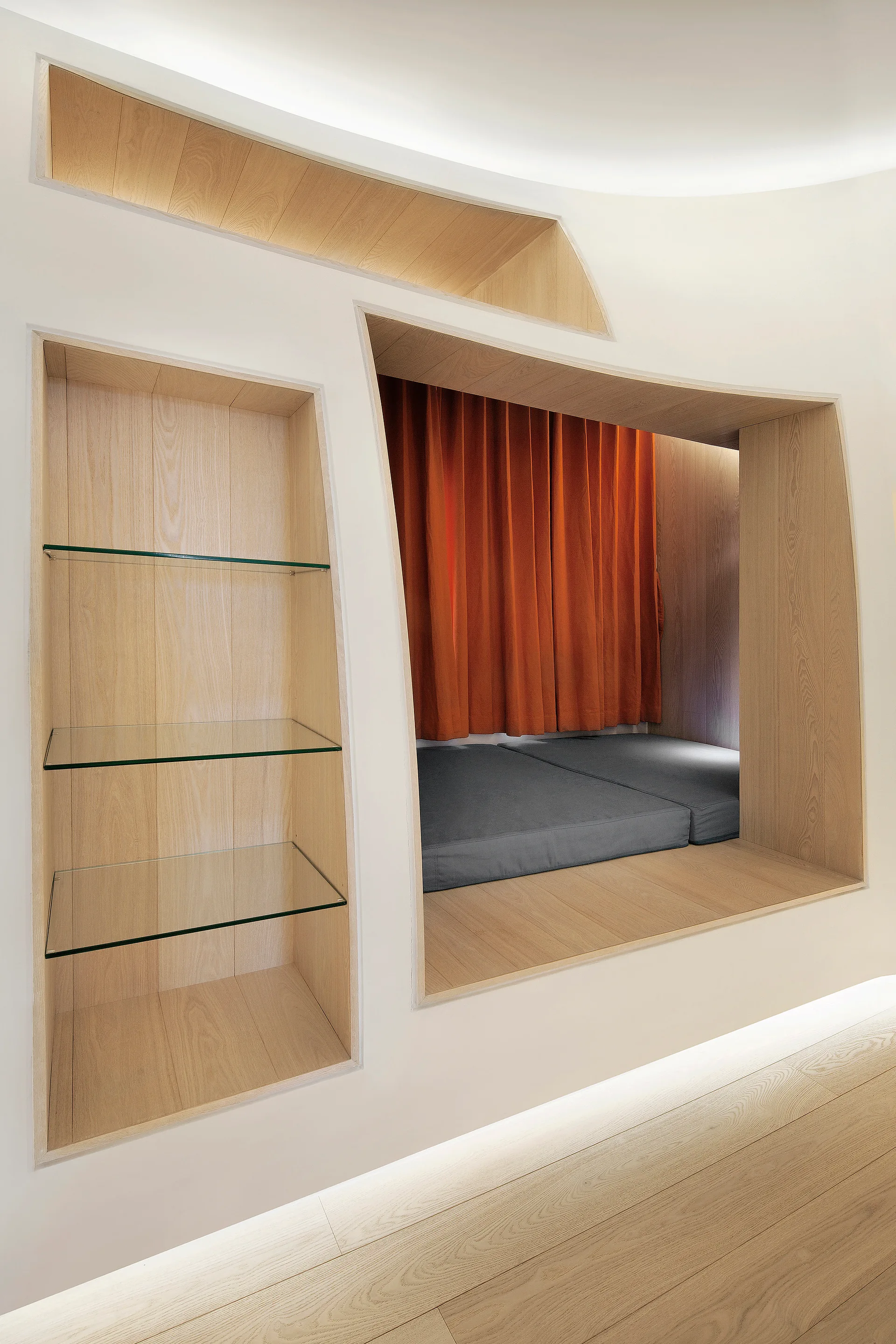 A custom light wood built-in unit features a recessed bed with grey mattresses and orange curtains, alongside open shelving with glass panels.