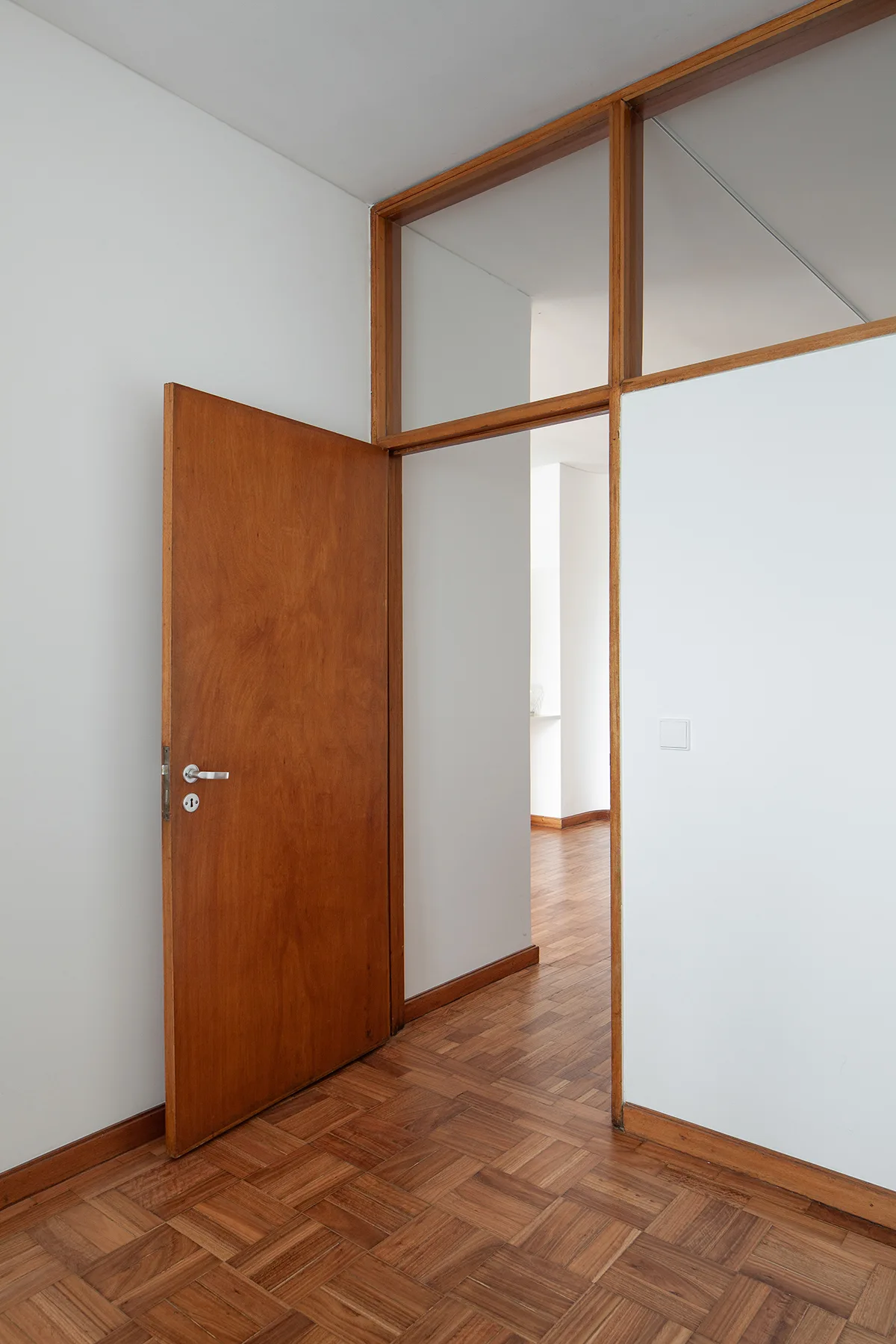 An open wooden door stands in a white hallway featuring parquet flooring and a wood-framed transom window above the doorway.
