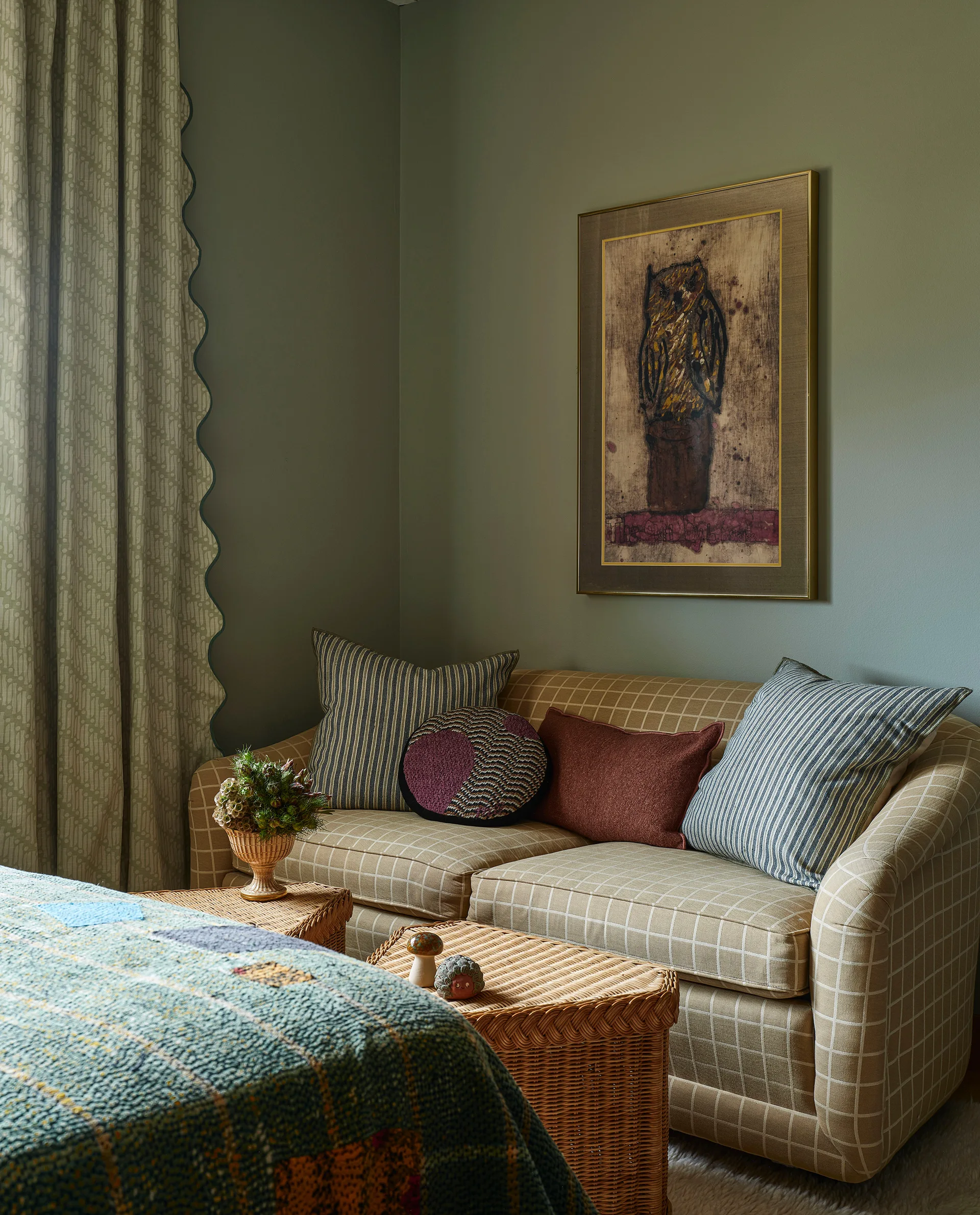 A sage green bedroom corner features a beige checkered sofa with accent pillows next to a wicker table and scalloped curtains.