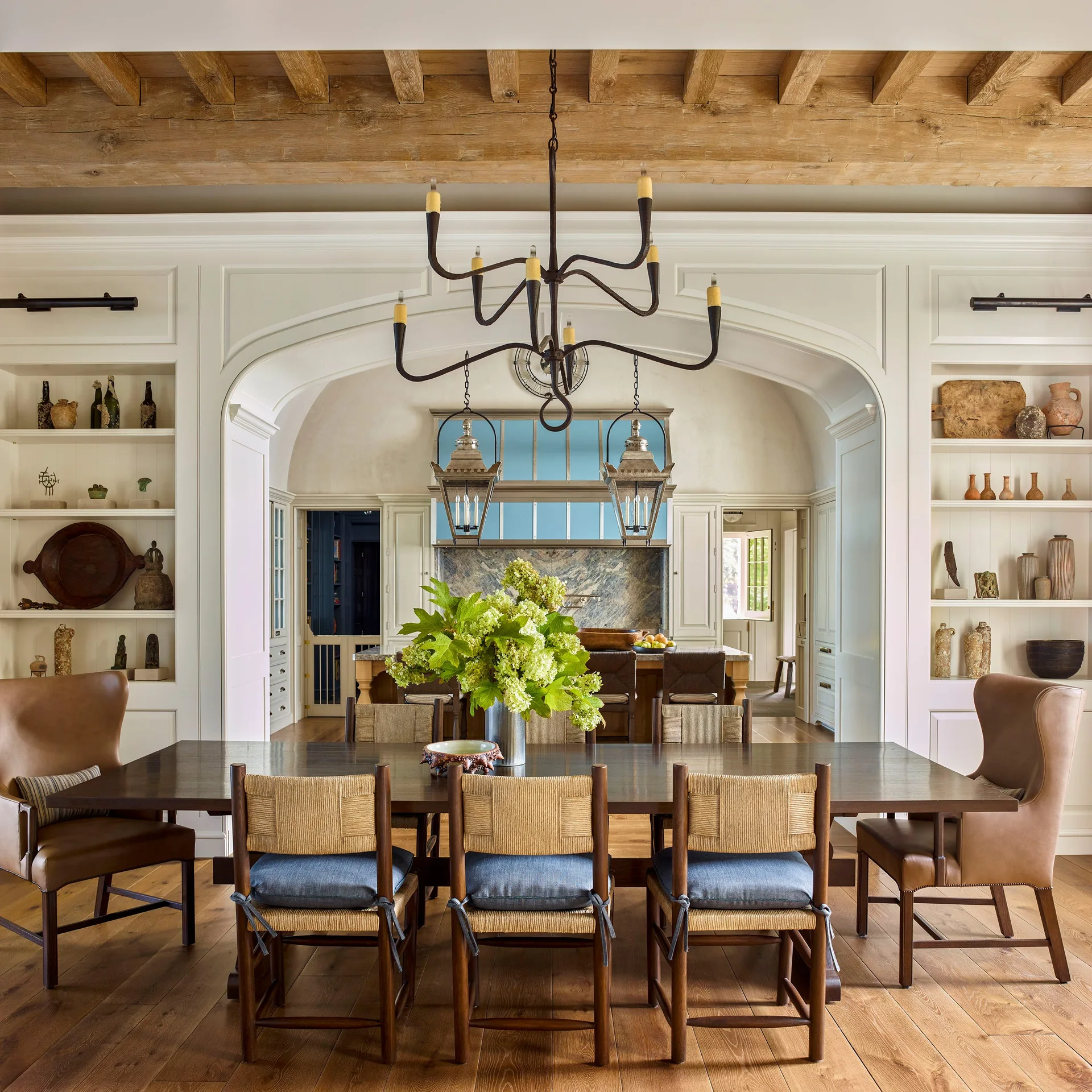A rustic dining room features a dark wood dining table, woven chairs, and an arched entryway leading into a kitchen with blue accents.