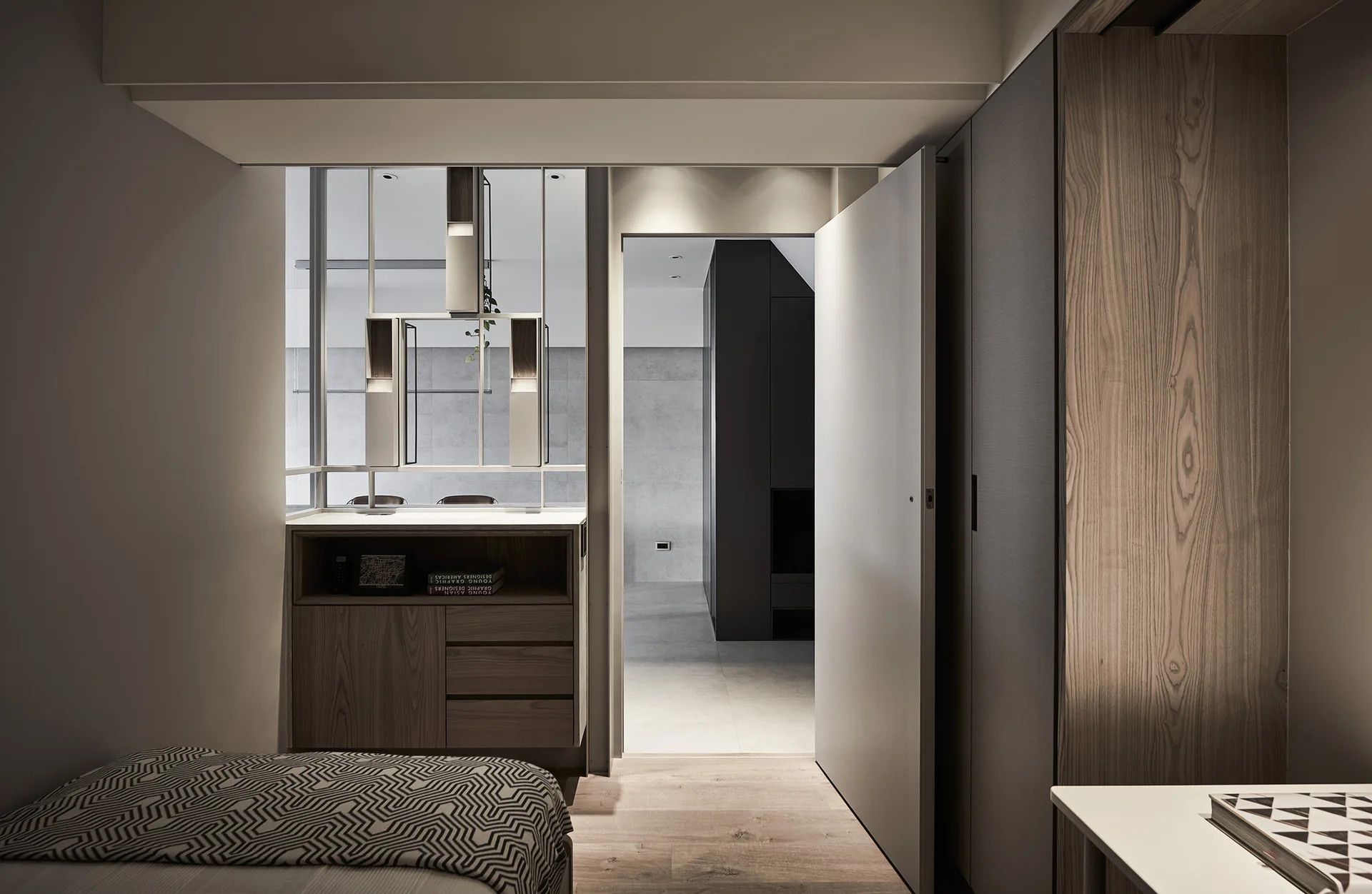 A modern bedroom features a bed with a geometric cover, built-in wood cabinets, and an open doorway looking into a minimalist adjoining room.