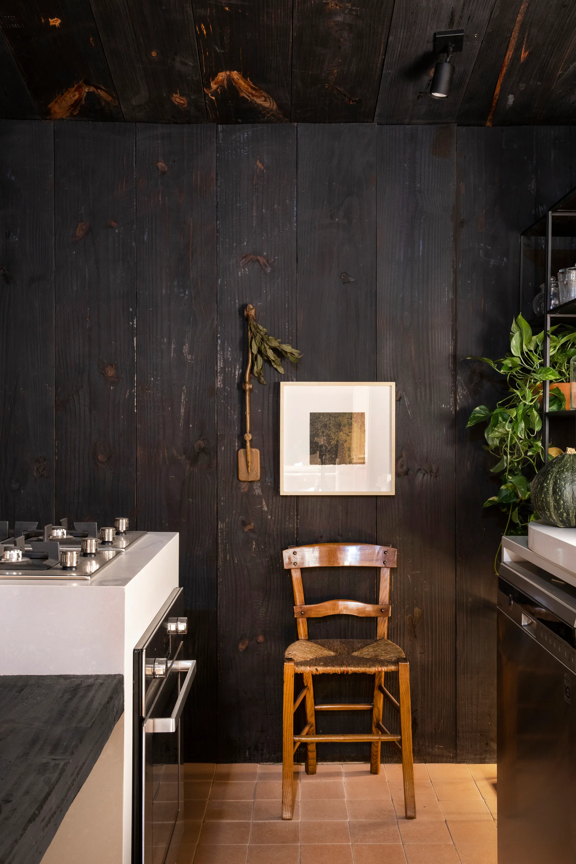 A kitchen featuring dark vertical wood paneling on the walls, terracotta floor tiles, a wooden chair, and stainless steel appliances.