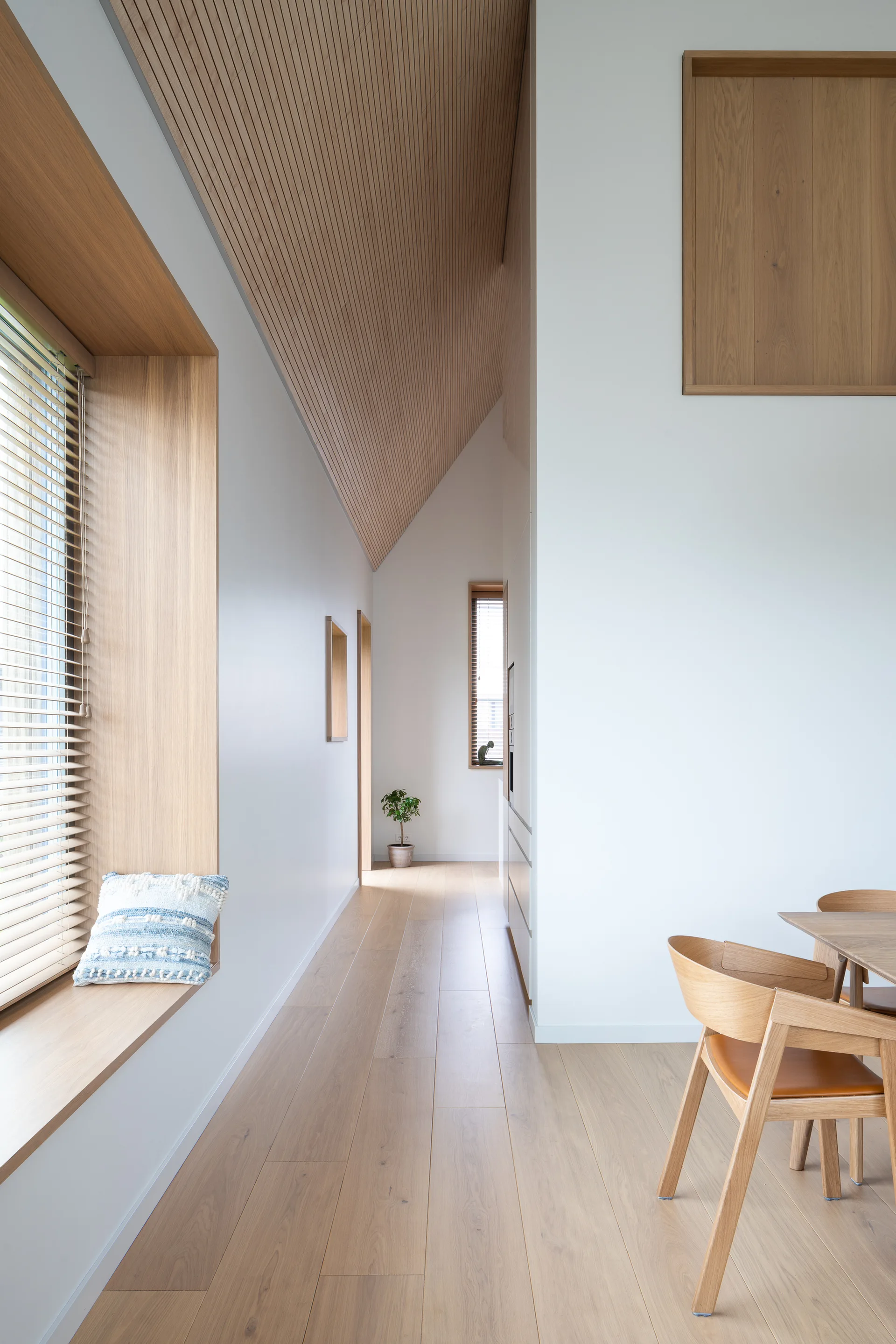 A modern Scandinavian interior featuring a slatted wood ceiling, a built-in wooden window seat with a patterned pillow, and wooden dining chairs.