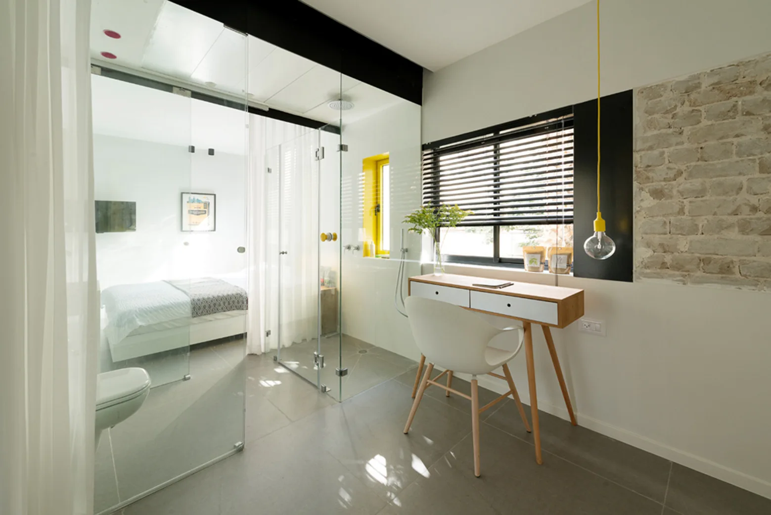 A modern bedroom suite features a glass-enclosed bathroom alongside a slim wooden desk, white chair, and an exposed brick wall accent.