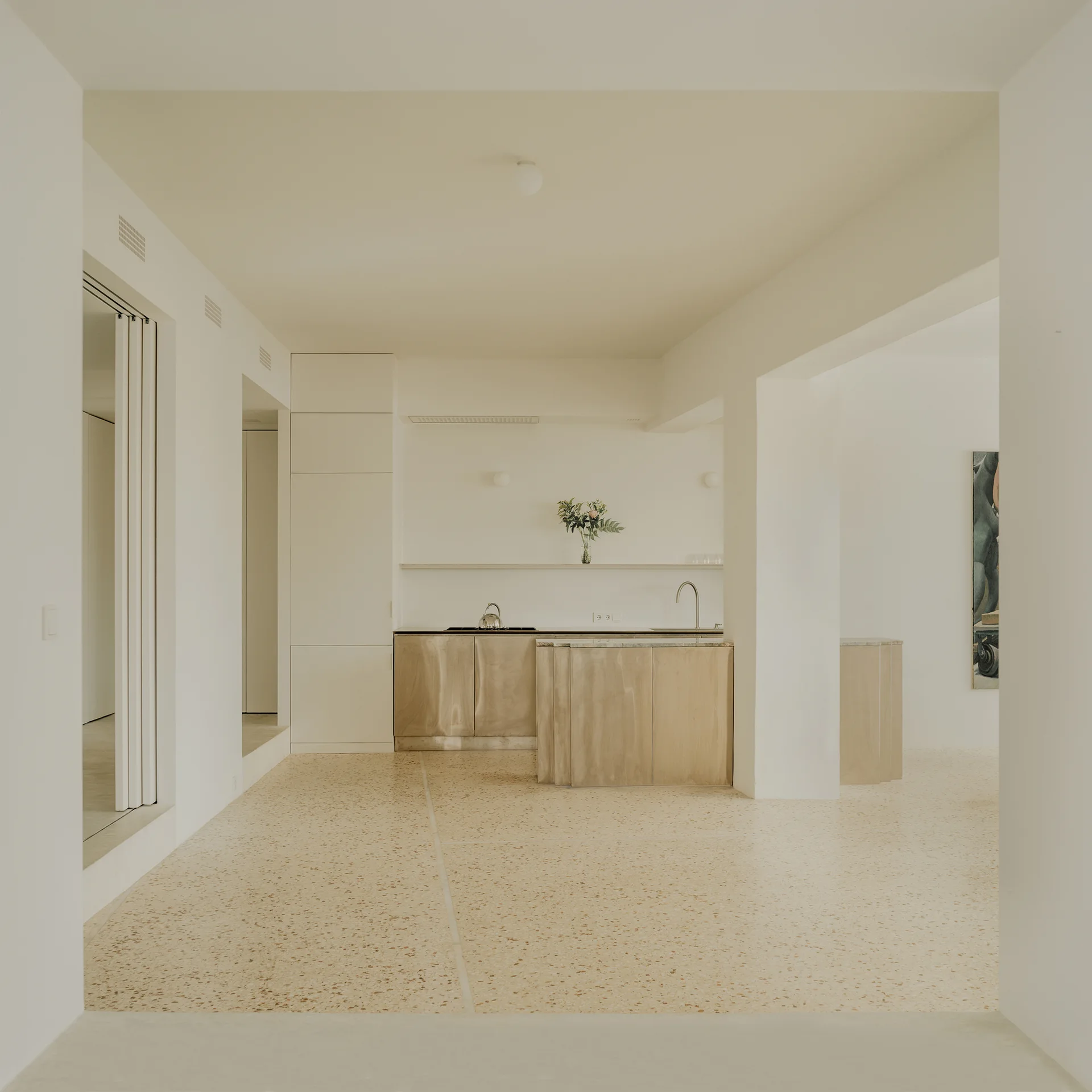 A minimalist kitchen featuring light wood lower cabinetry, a metal faucet, and a large expanse of speckled beige terrazzo flooring under an off-white ceiling.