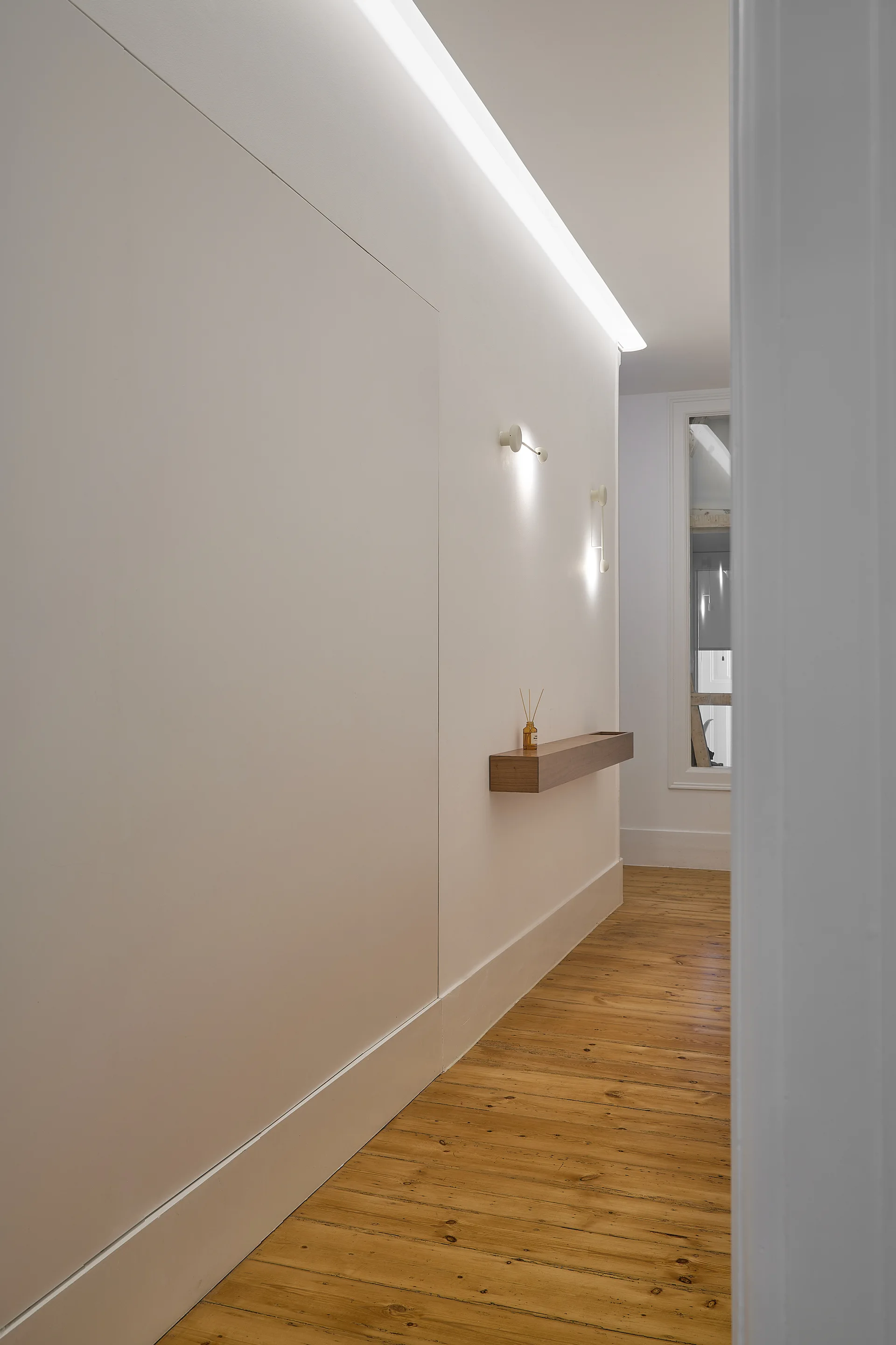 A minimalist white hallway featuring natural wood plank flooring, a small floating wooden shelf, and soft cove lighting along the ceiling.