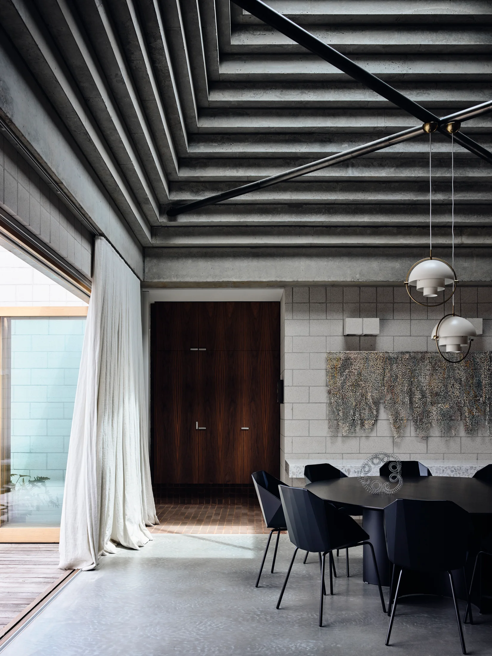 A modern dining room features a round black table, matching chairs, a tiered concrete ceiling, concrete block walls, and soft white curtains.