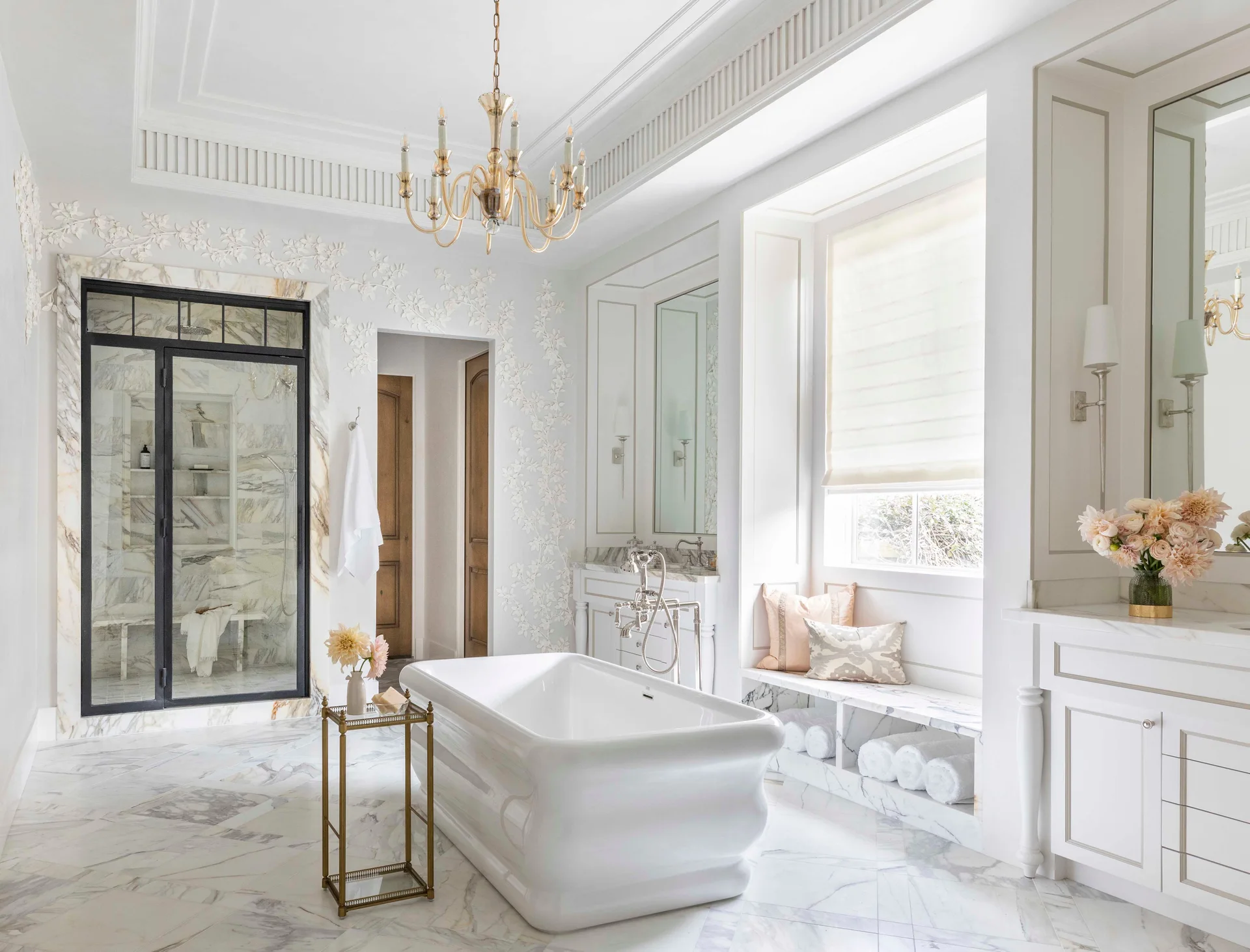 A white marble bathroom features a freestanding bathtub, a black-framed glass shower, a gold chandelier, and floral relief details on the walls.