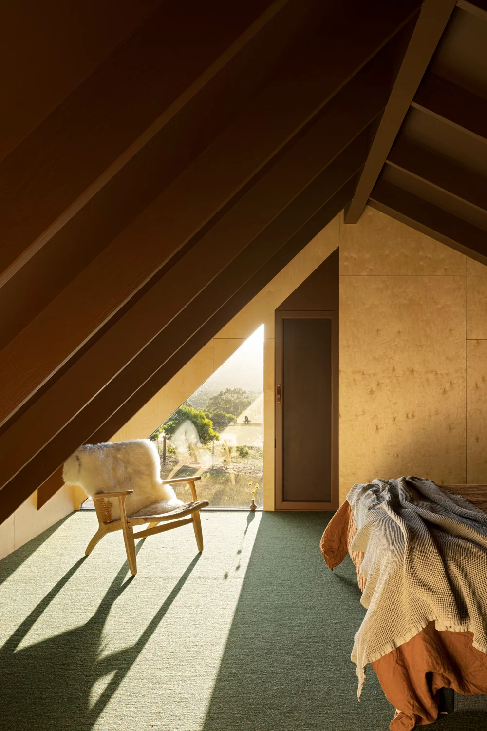 An attic bedroom featuring a steep sloped wood ceiling, a triangular window with sunlight streaming in, a wooden armchair, and green carpet.