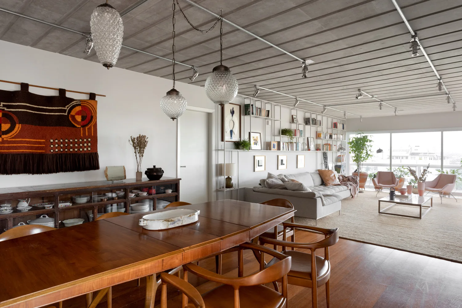 An open-concept living and dining space featuring a wood dining table, a grey sofa, white metal shelving, and a textured concrete ceiling.