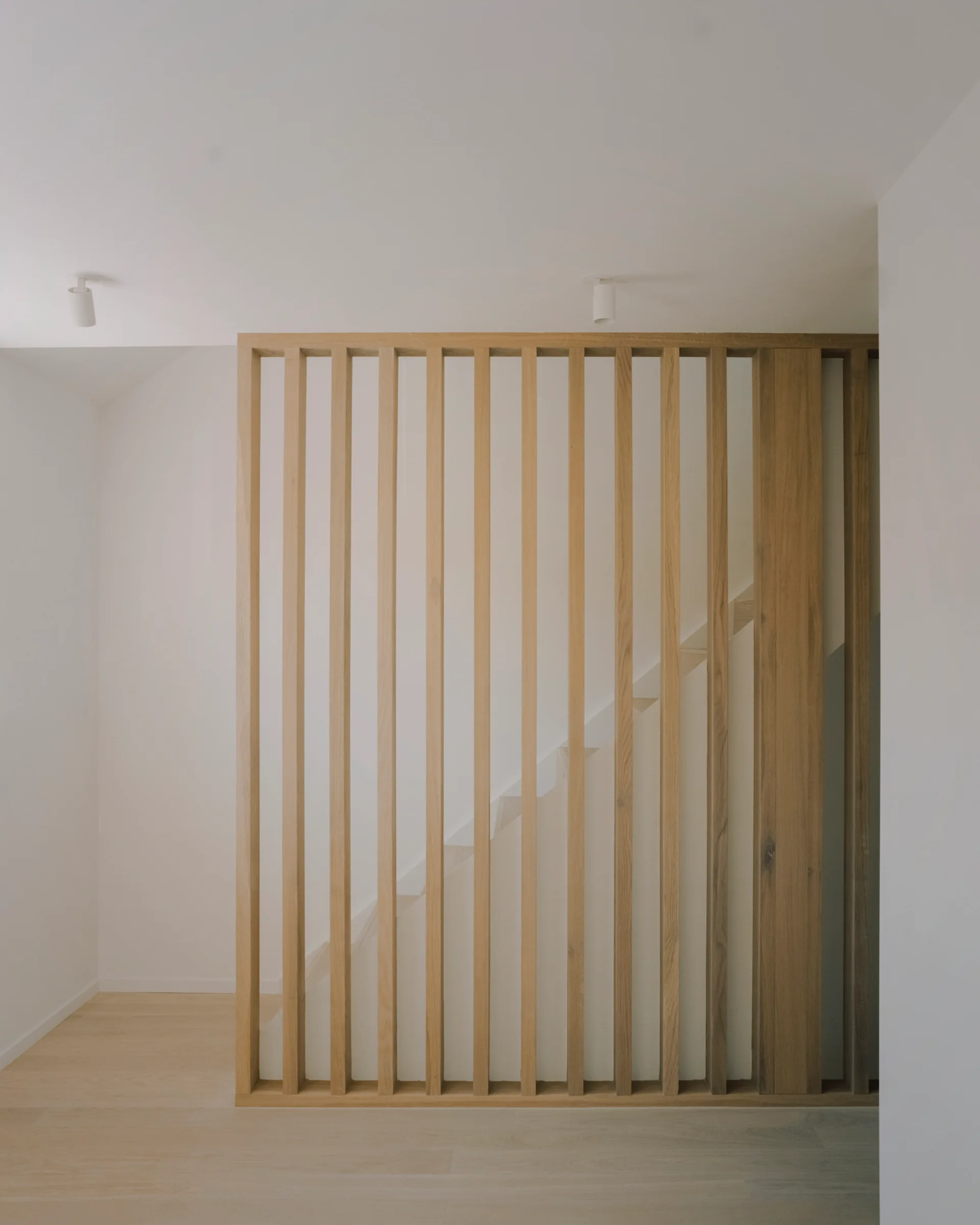 A light wood vertical slatted screen partition separates a minimalist white hallway from a staircase with matching light wood flooring.