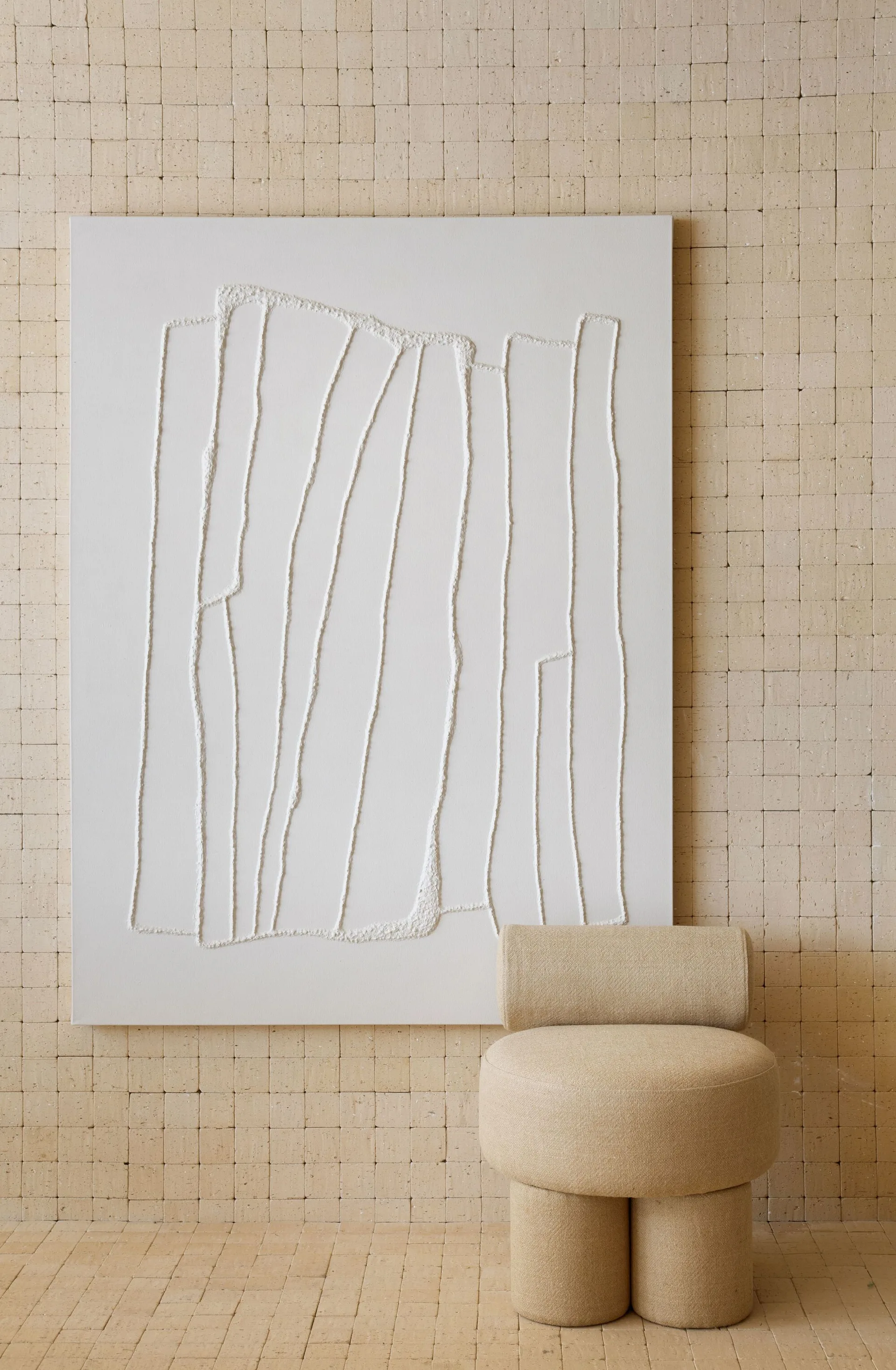 An upholstered beige sculptural stool stands in front of a large textured white canvas on a wall covered in small square tiles.
