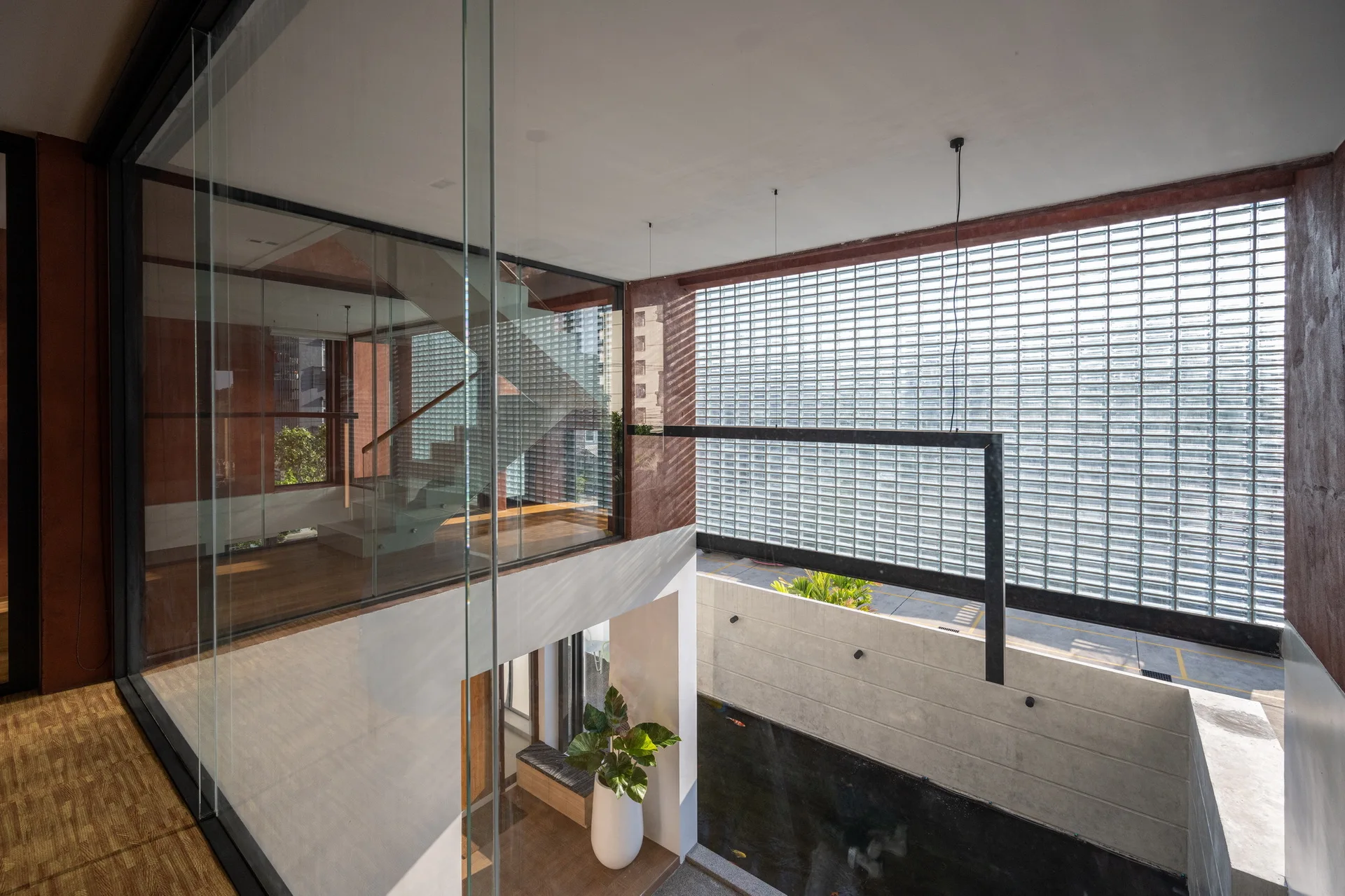 A modern multi-level interior featuring a glass-enclosed staircase overlooking an internal courtyard with a water pond and a large glass block wall.