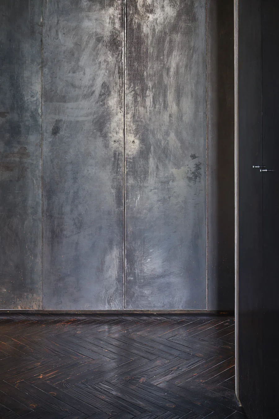 Tall patinated gray cabinet doors stand above a dark wood floor laid in a herringbone pattern.