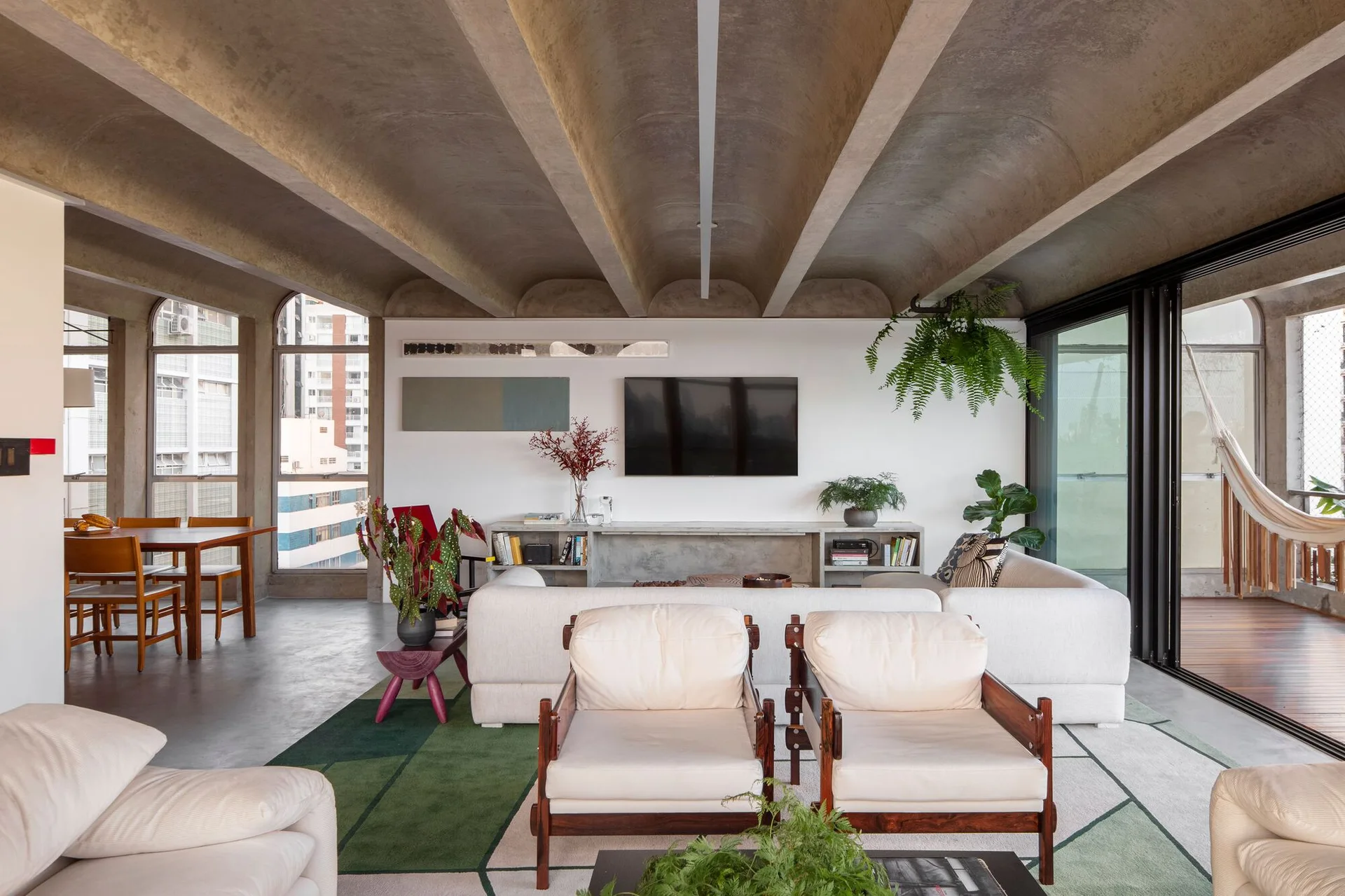 A modern living room features a curved concrete vaulted ceiling, a cream sofa with wooden armchairs, and a large green geometric rug.