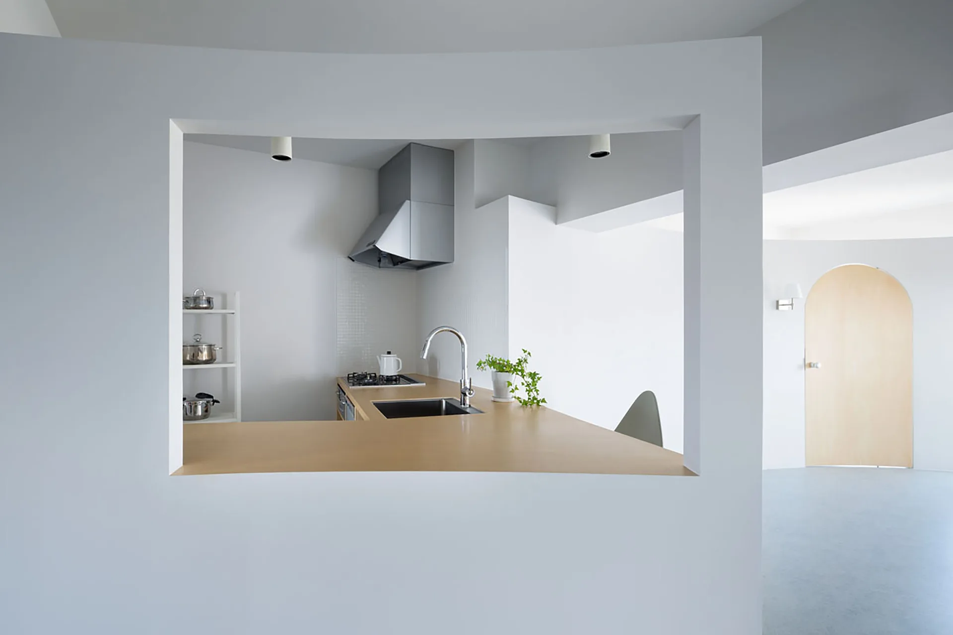 A minimalist kitchen with a light wood countertop is visible through a large rectangular wall cutout next to a light wood arched doorway.