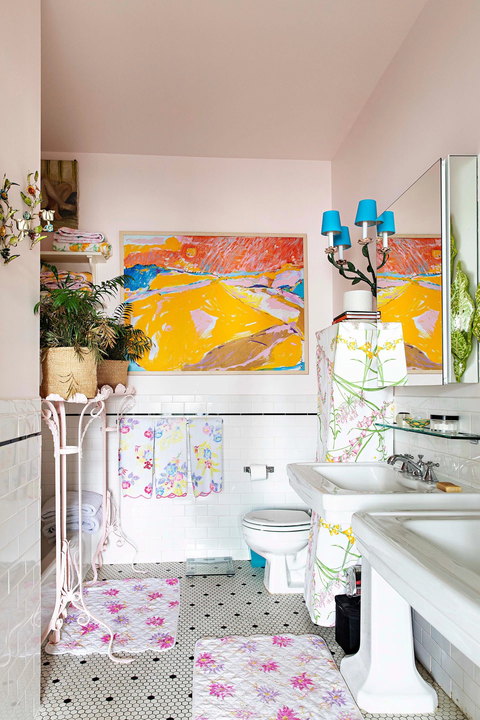 A pink bathroom features white subway tiles, a black-and-white mosaic tile floor, floral bath mats, and a large colorful painting on the wall.