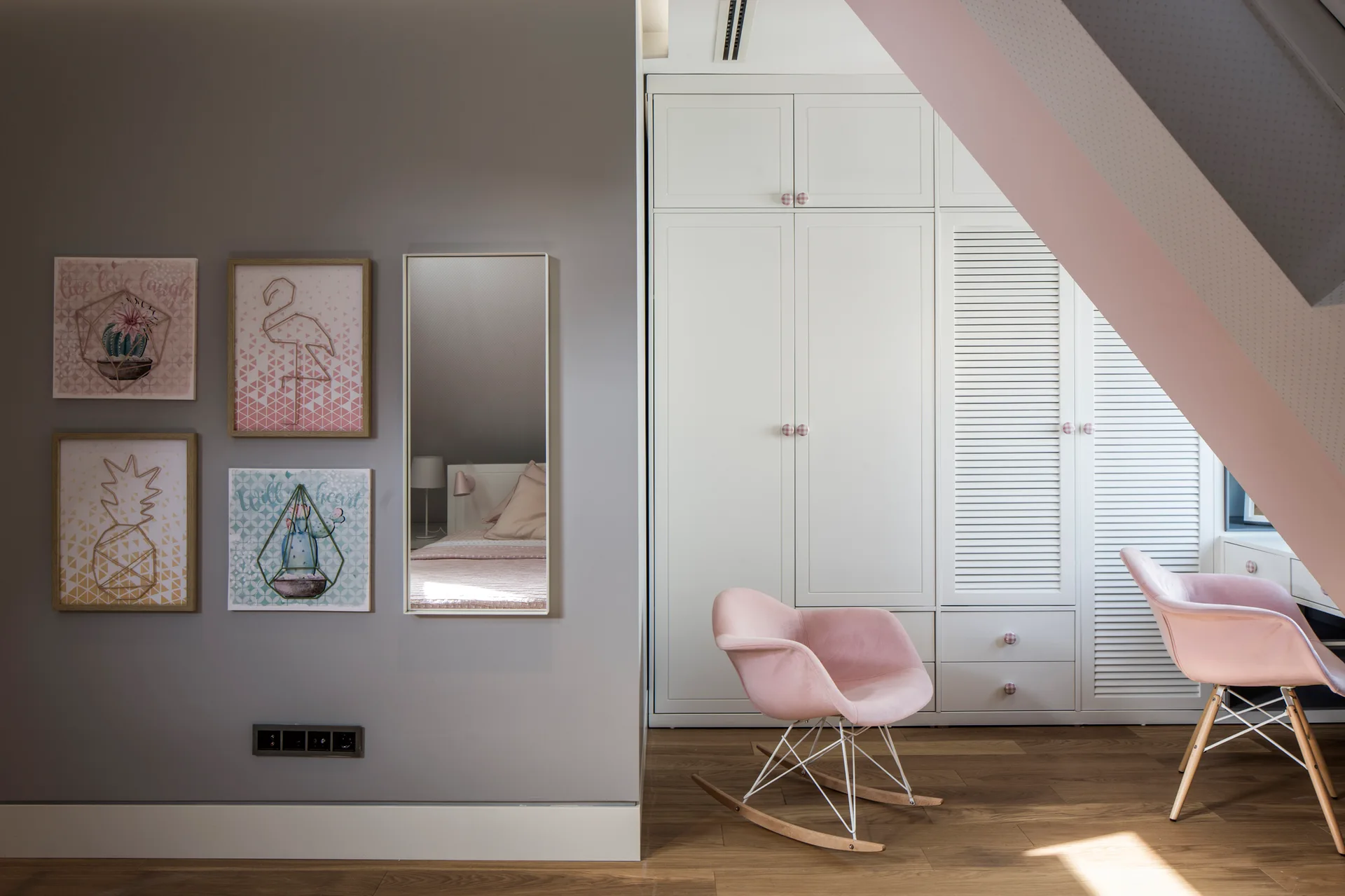 A children's bedroom features a gray accent wall with four framed art prints, a tall white built-in wardrobe, and two pink modern chairs.