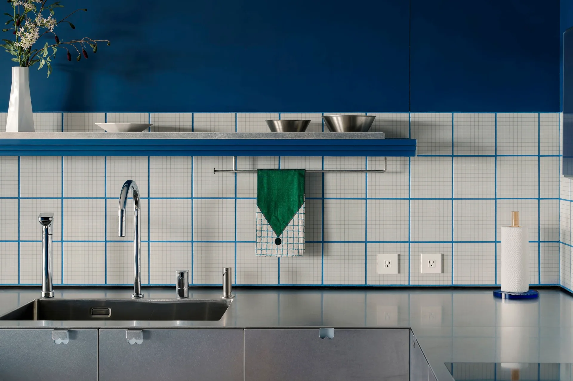 A modern kitchen features a stainless steel countertop and sink set against a white square tile backsplash with blue grout lines and a blue upper wall.