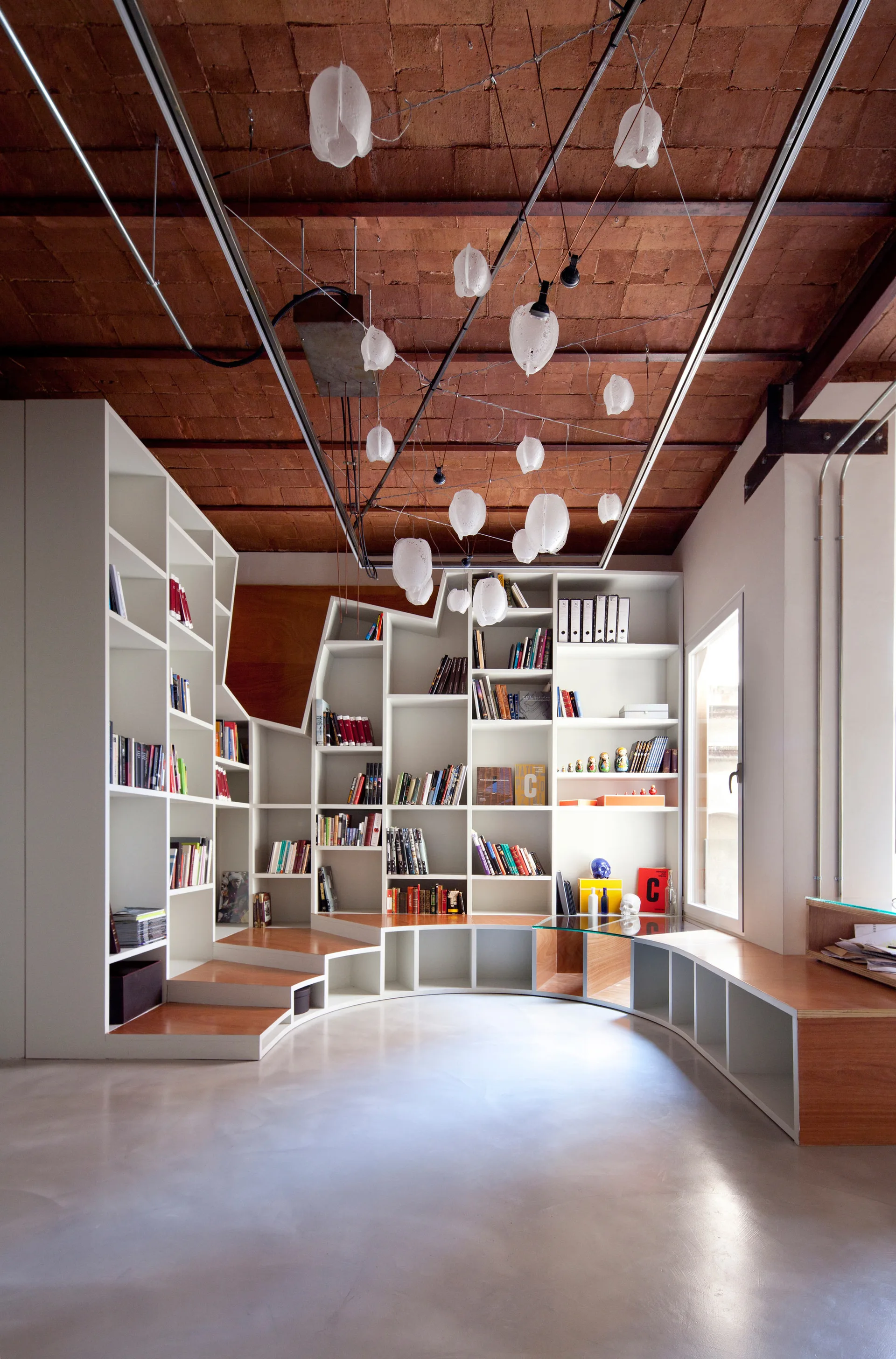 A large white custom bookshelf with integrated wooden steps curves along a wall under an exposed brick ceiling with hanging pendant lights.