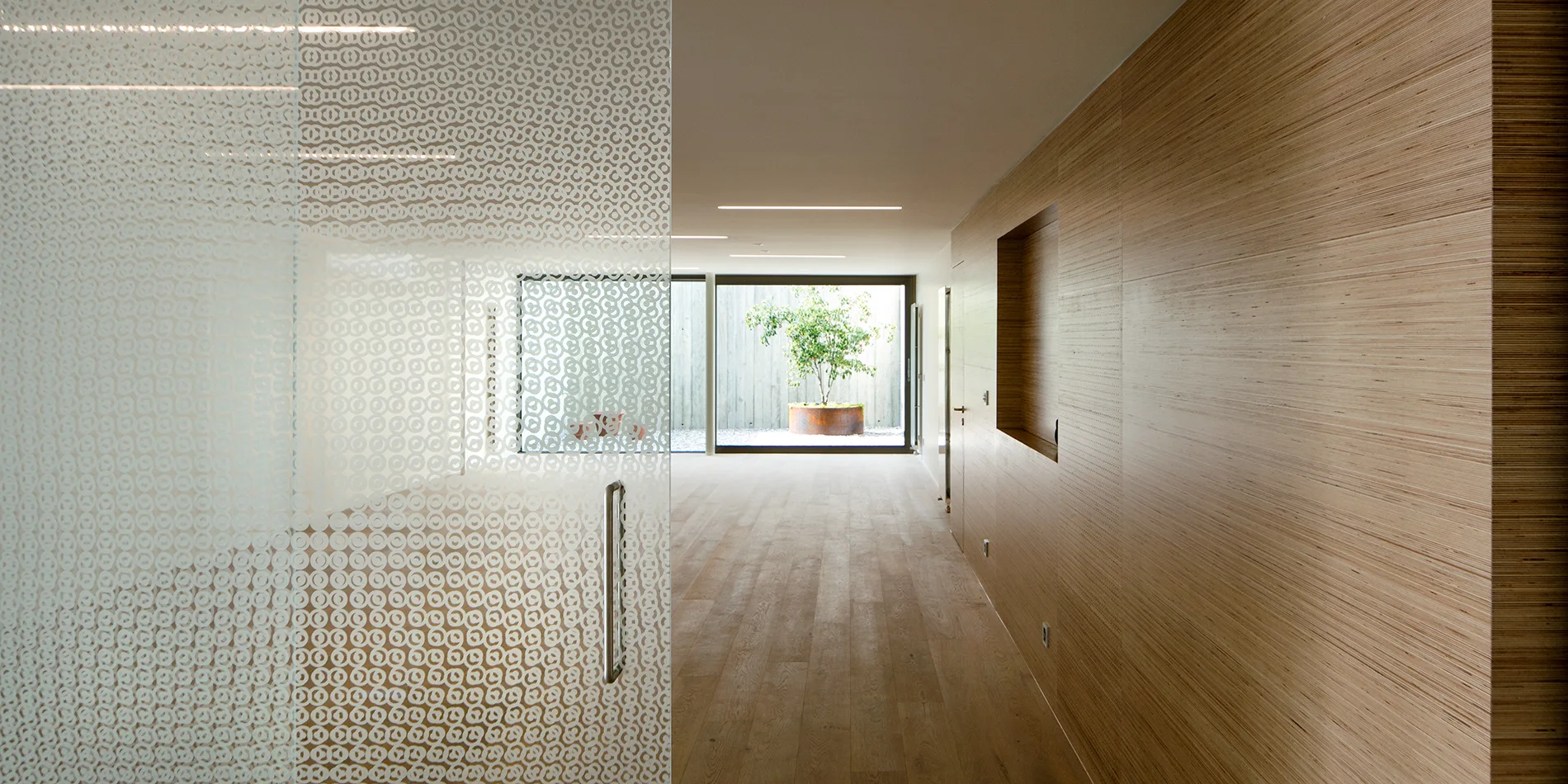 A minimalist hallway features horizontal wood wall paneling, linear ceiling lights, and a patterned frosted glass partition leading to an outdoor courtyard.