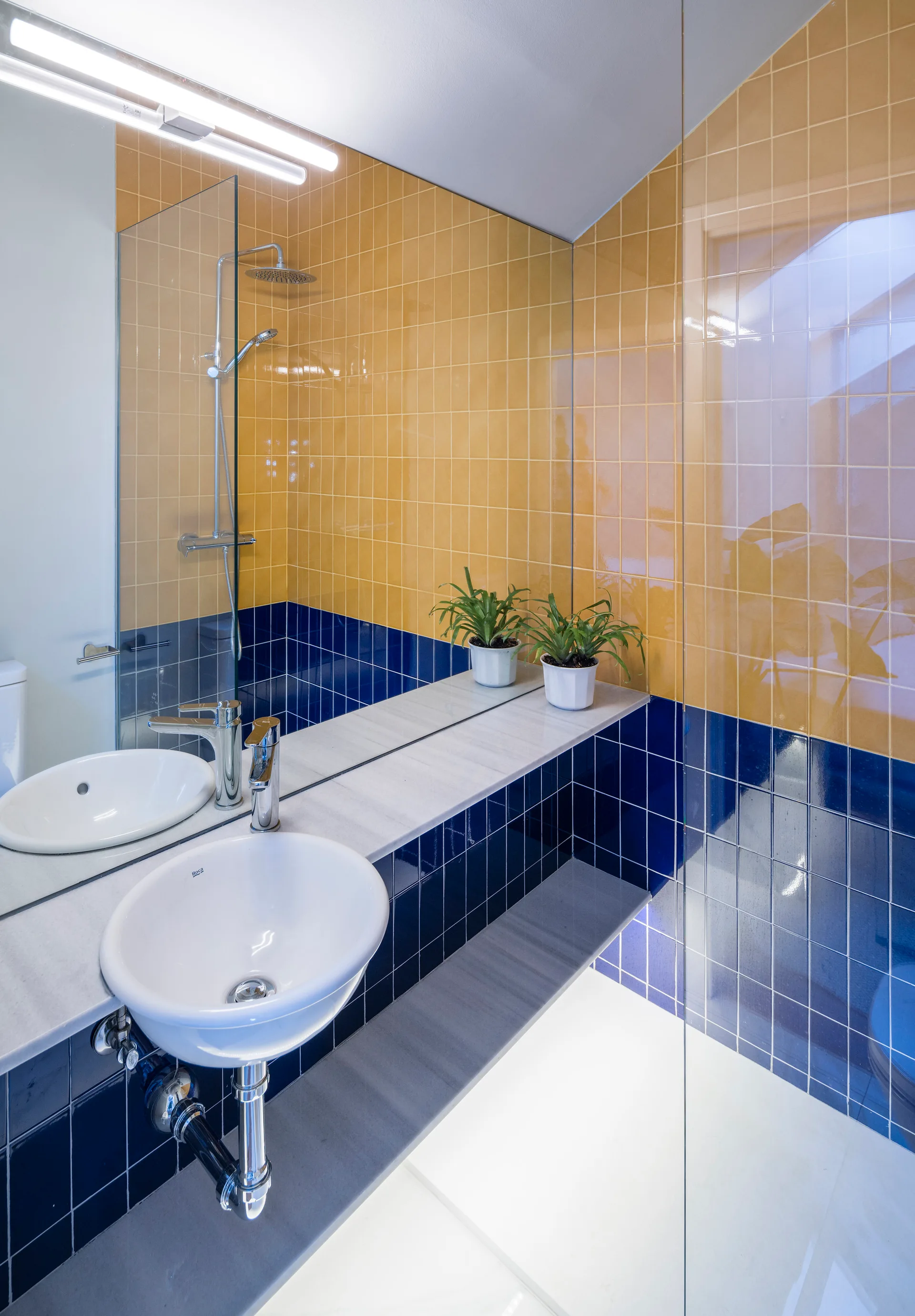 A contemporary bathroom features a wall with yellow and navy blue square tiles, a white basin sink, a large mirror, and a glass shower enclosure.