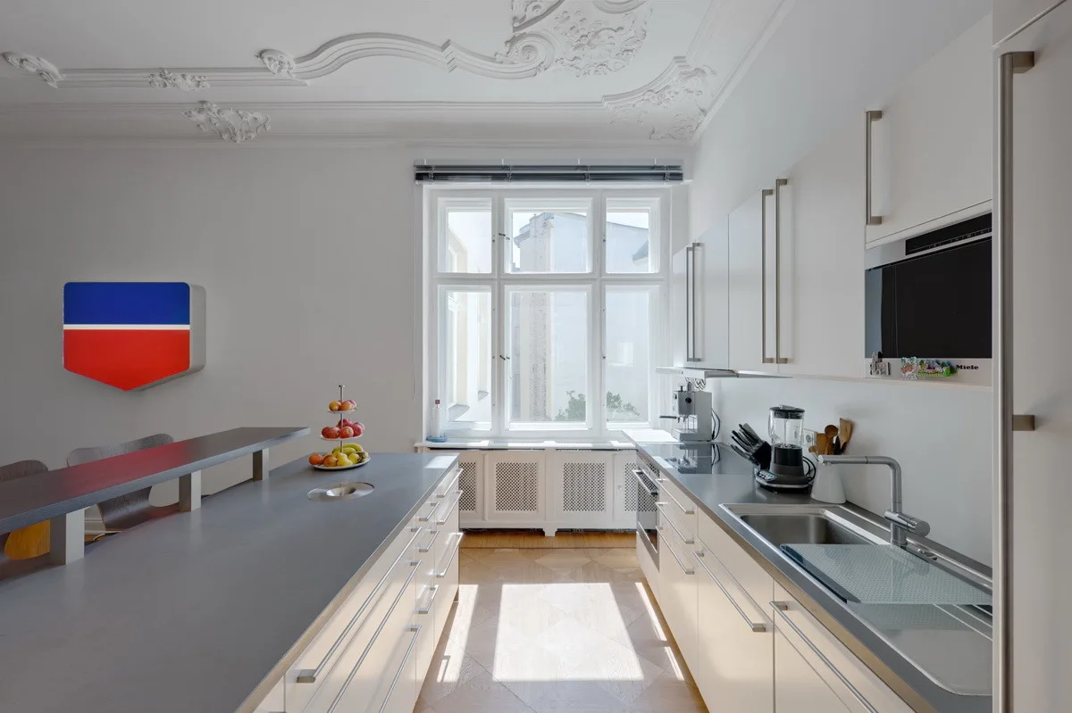 A bright galley kitchen features minimalist white cabinets, grey countertops, a central island, and ornate historic ceiling molding.