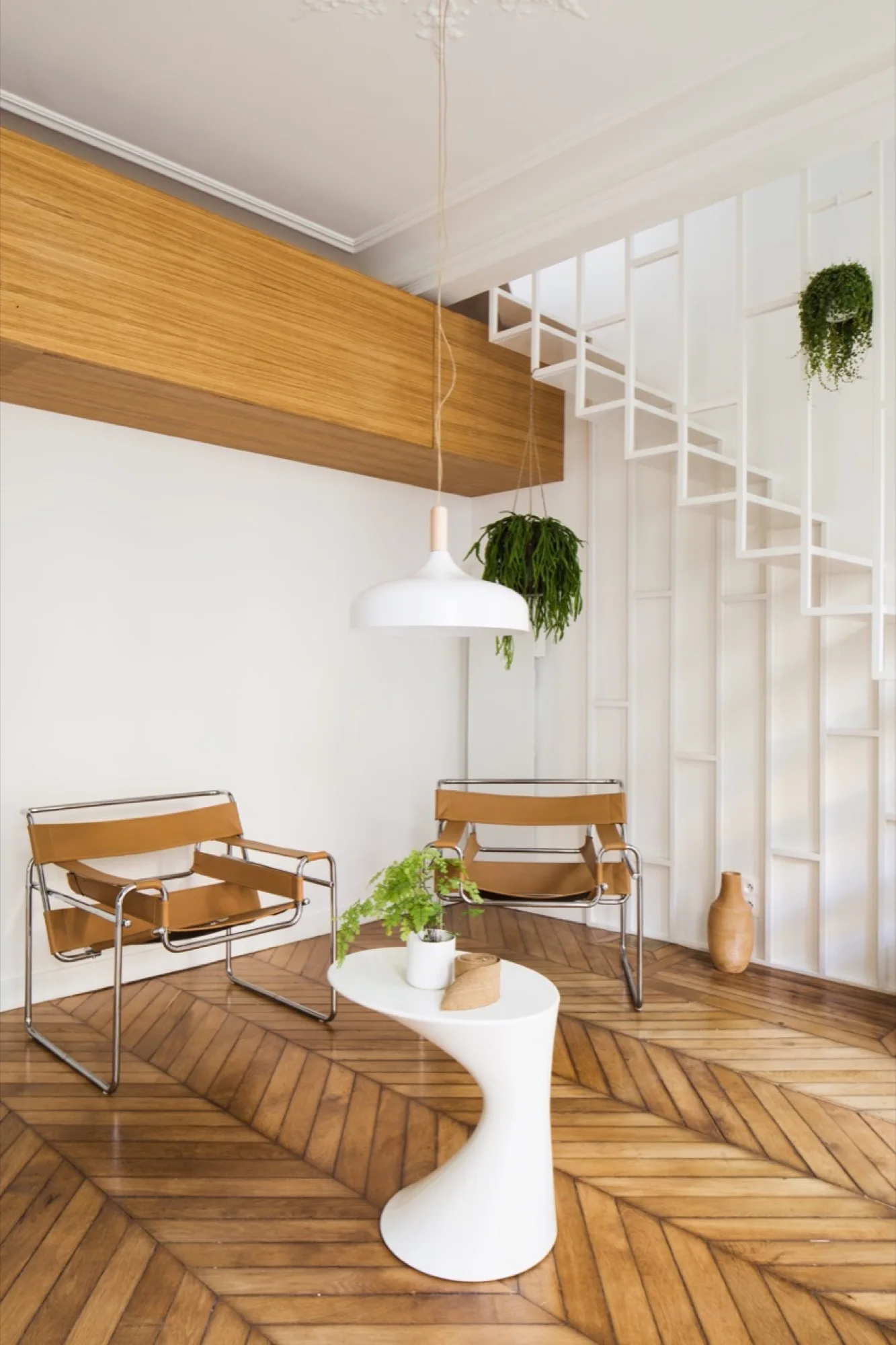 Two tan leather strap armchairs sit on a chevron wood floor in a white minimalist living area next to an open geometric staircase.