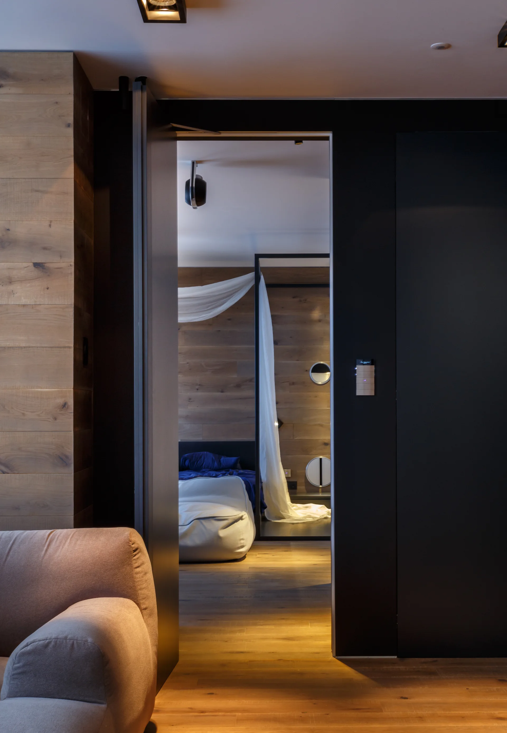 A view through a matte black door frame reveals a warm bedroom featuring horizontal wood paneling, a bed with blue bedding, and draped sheer white fabric.