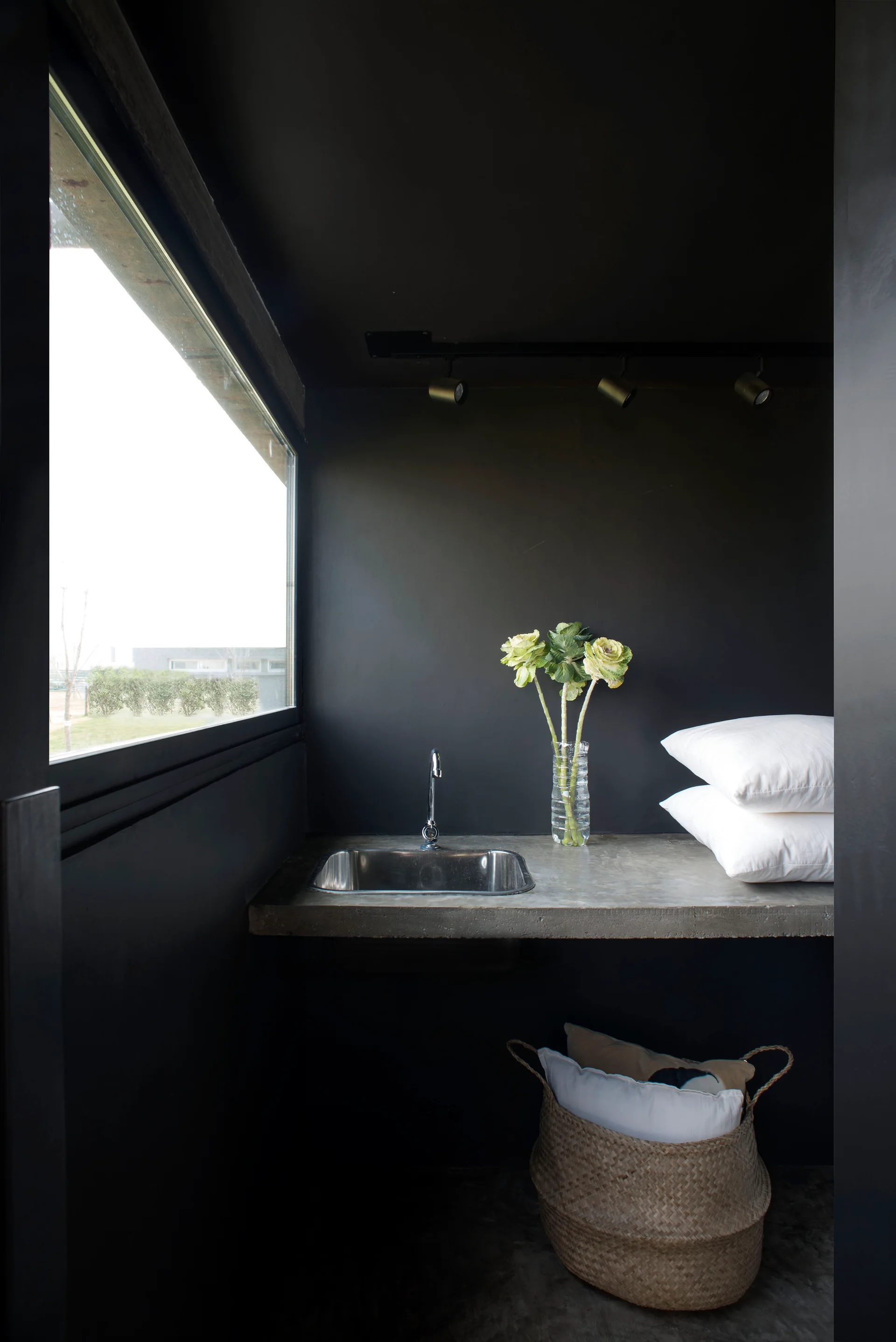 A concrete countertop with an integrated metal sink sits against matte black walls under a large window, next to stacked white pillows.