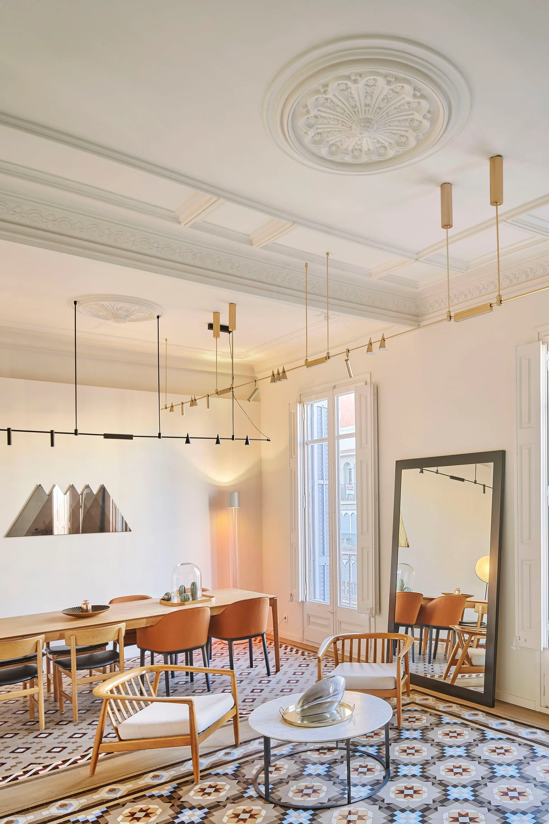 A bright dining room features a light wood table with orange chairs, geometric patterned tile flooring, and highly decorative white ceiling moldings.