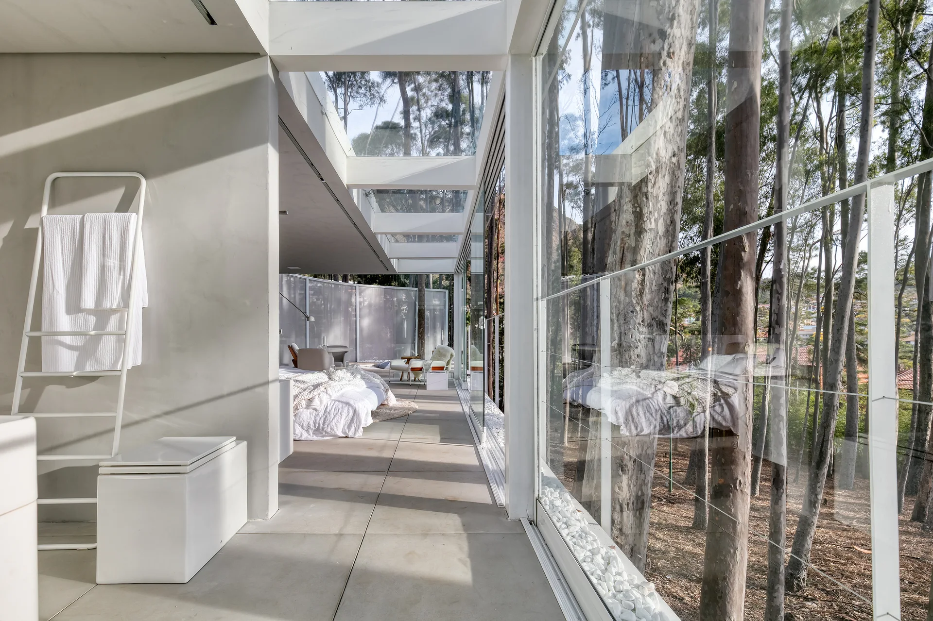 A minimalist modern bedroom corridor featuring floor-to-ceiling glass walls, grey concrete-style floor tiles, and a white towel ladder.