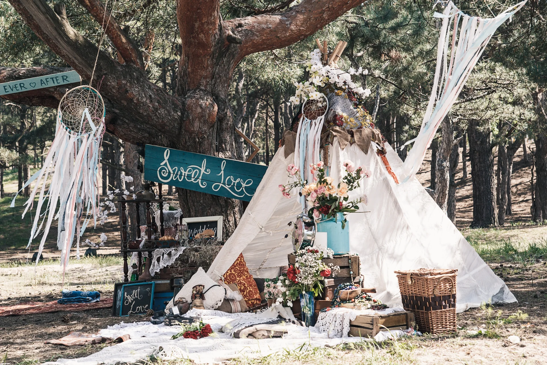 A white canvas teepee decorated with dreamcatchers and floral arrangements under a pine tree in a forest setting.
