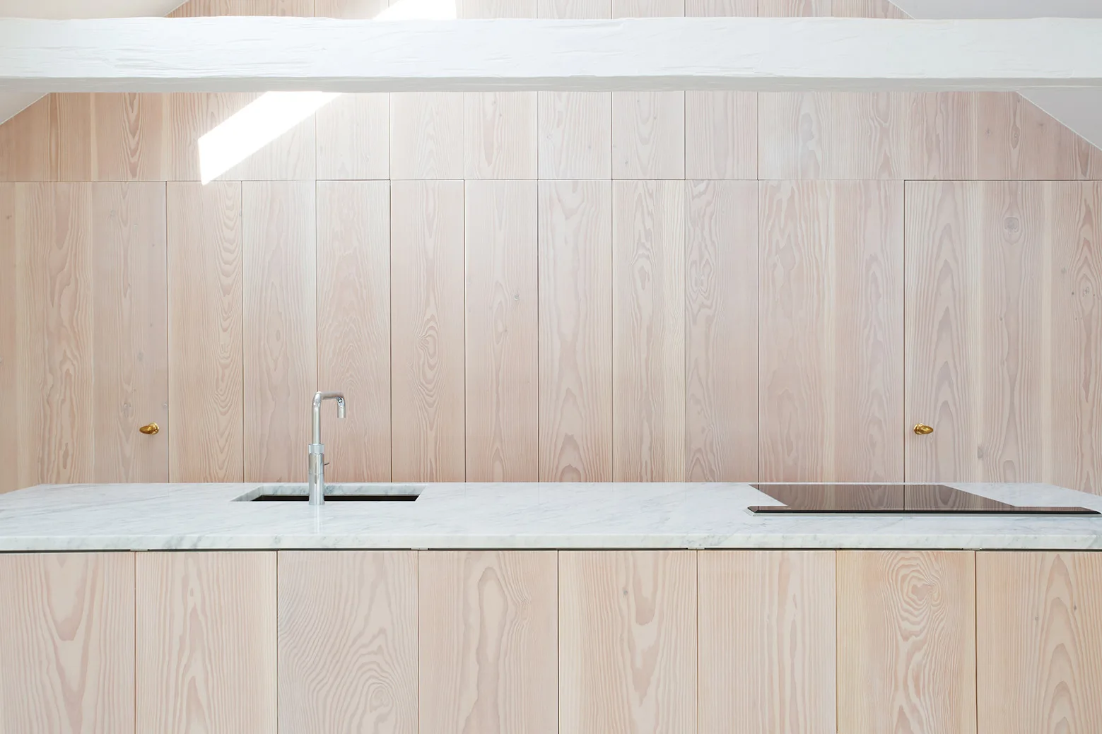 A minimalist kitchen features a white marble island with a silver faucet and integrated cooktop against vertical light wood paneling.