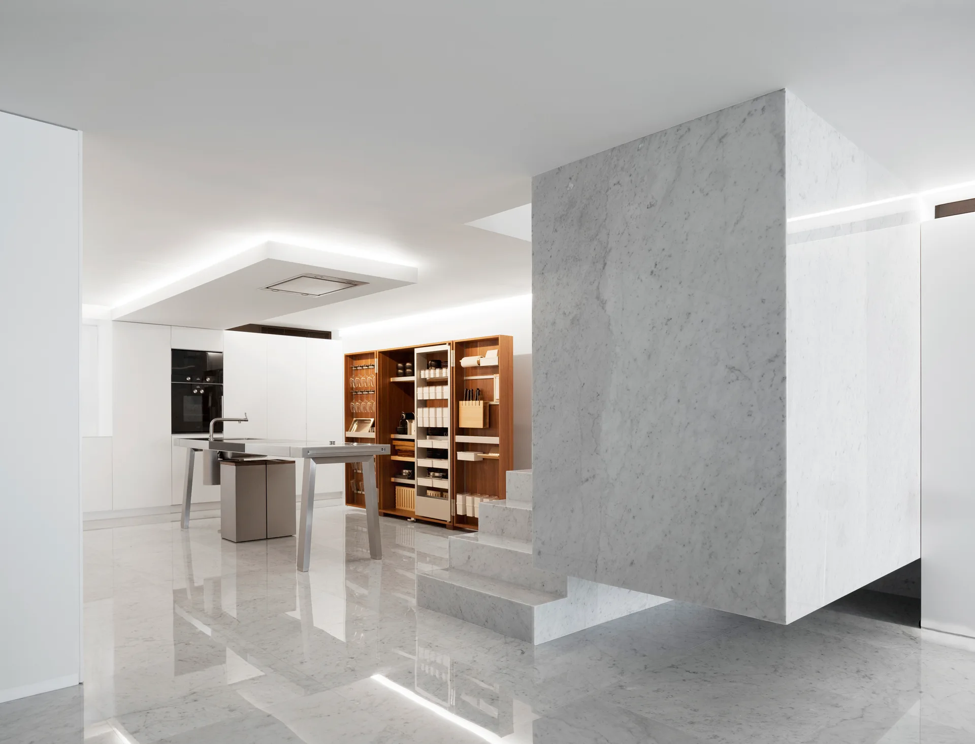A minimalist white kitchen features a large floating marble wall piece above stairs, a stainless steel island, and open wood shelving.