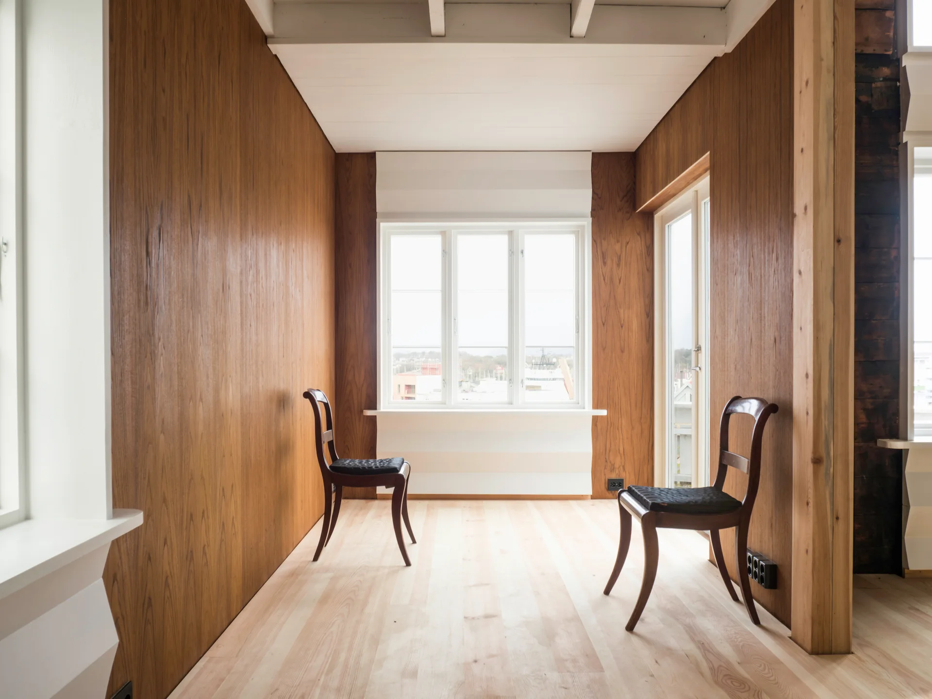 A minimalist room features vertical warm wood wall paneling, light wood plank floors, a white beamed ceiling, and two dark wooden chairs.