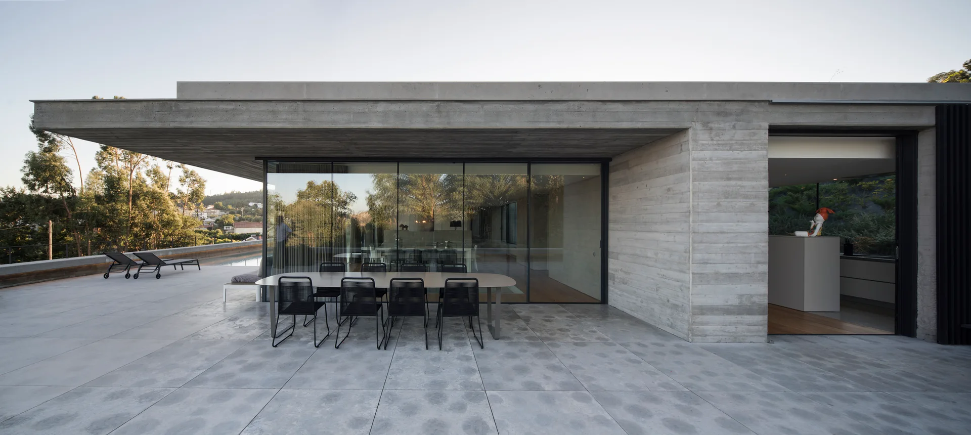 A minimalist outdoor concrete terrace features a black dining set beneath a large cantilevered roof overhang next to glass walls.