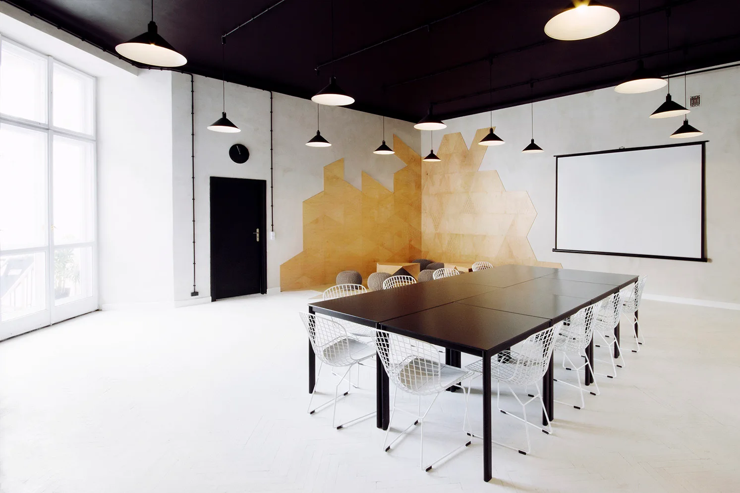 A long black conference table is surrounded by white wire chairs under a black painted ceiling with pendant lights and geometric wood wall panels.
