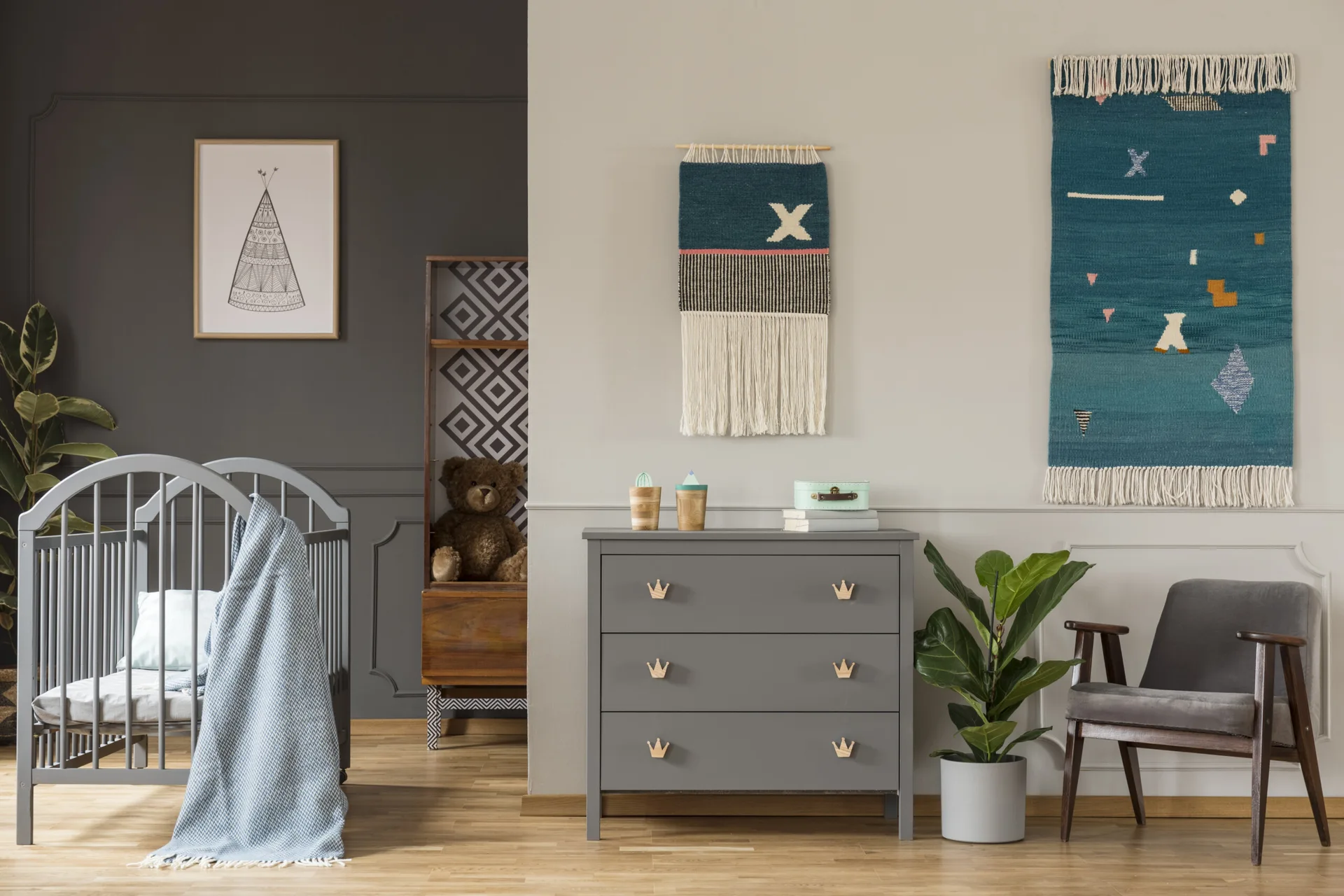 A modern nursery with a grey wooden crib, a matching three-drawer dresser, a grey armchair, and colorful woven wall hangings.